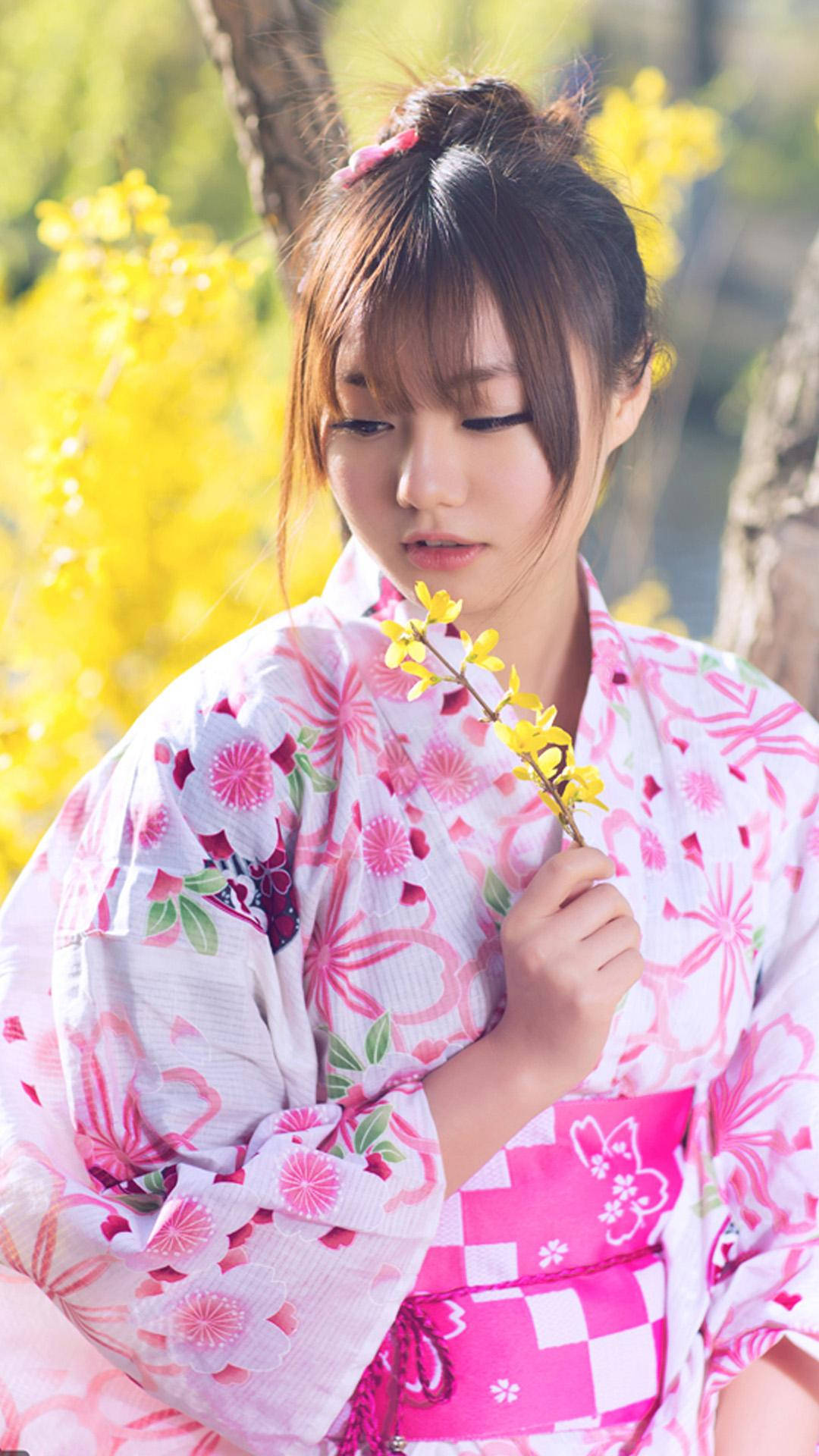 穿粉紅色和服的日本女孩 壁紙 — 一位穿著粉紅色花卉和服的年輕女子，在陽光明媚的春天背景下手持黃色花枝。