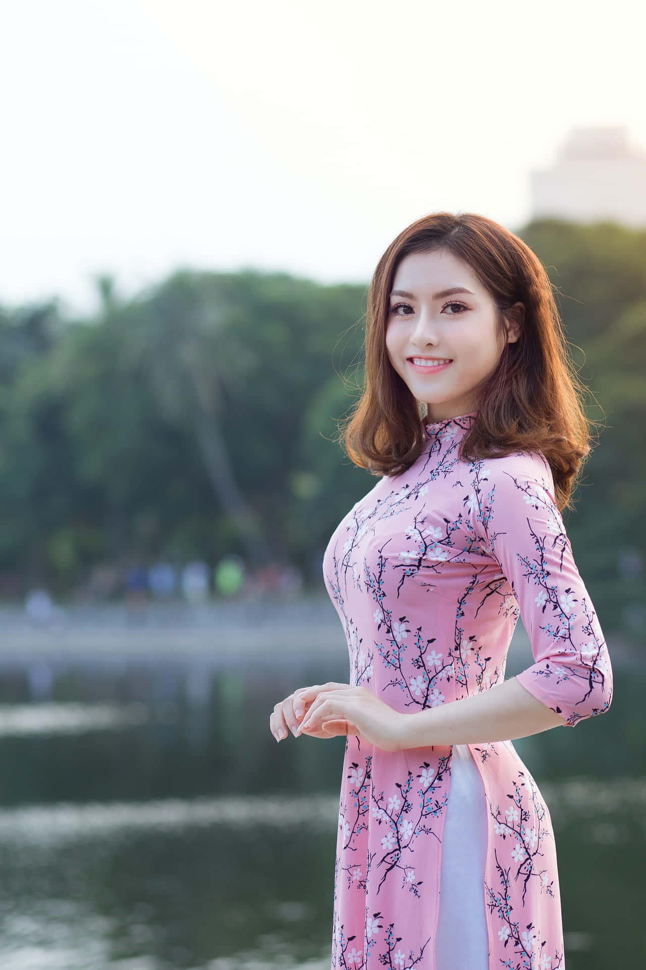 身穿粉紅奧黛的女子在湖邊微笑，背景有樹木。
