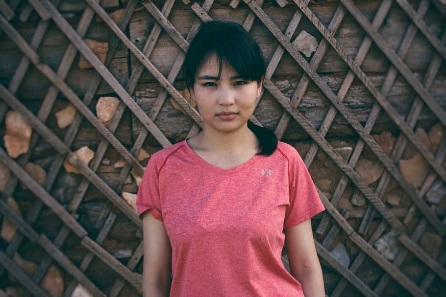 穿粉紅衣的蒙古少女 壁紙 — 一位身穿珊瑚色運動衫的年輕女子冷靜地站在帶有石頭裝飾的質樸木格柵牆前。