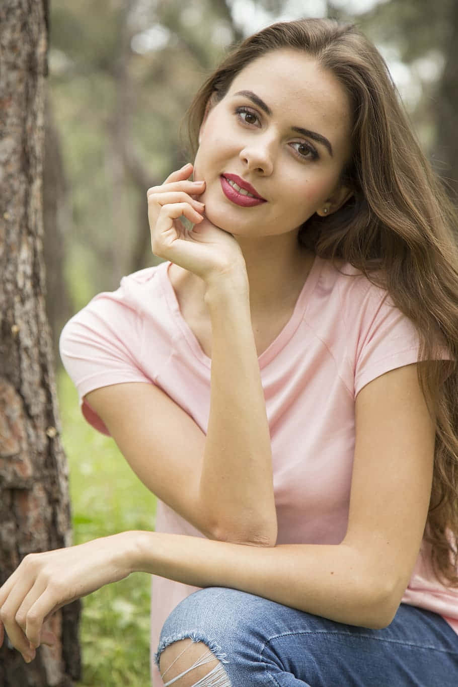 年輕女子身穿粉紅色襯衫和破洞牛仔褲靠著森林中的樹
