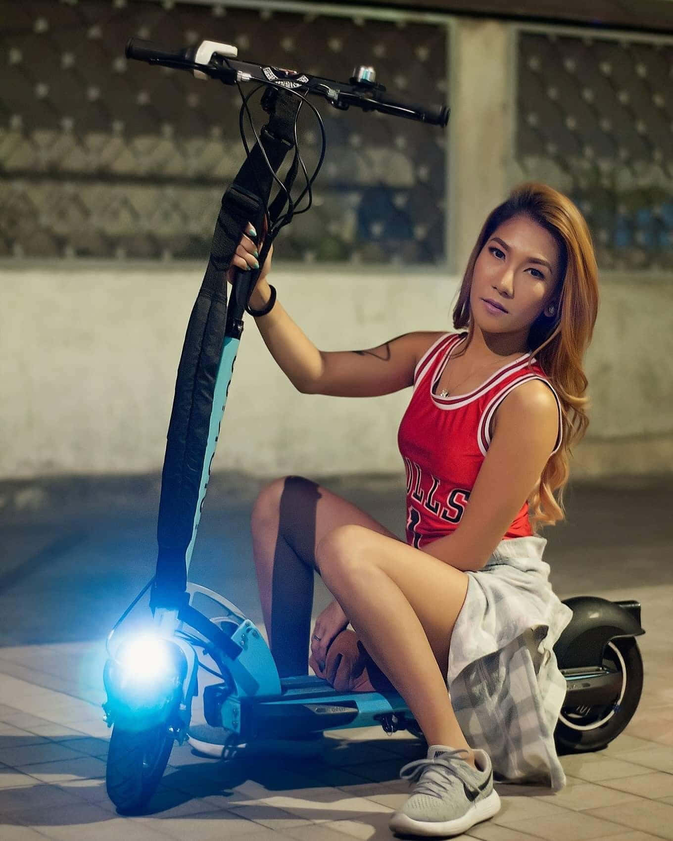 年輕女性身穿紅色球衣，夜晚坐在發光的藍色電動滑板車上。