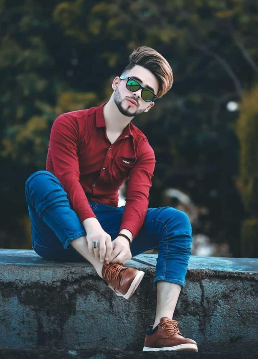 穿紅色Polo衫男子坐牆上 壁紙 — 一位時尚的年輕男子穿著紅色襯衫和藍色牛仔褲，坐在石頭邊緣調整他的棕色鞋子，同時戴著綠色太陽眼鏡。