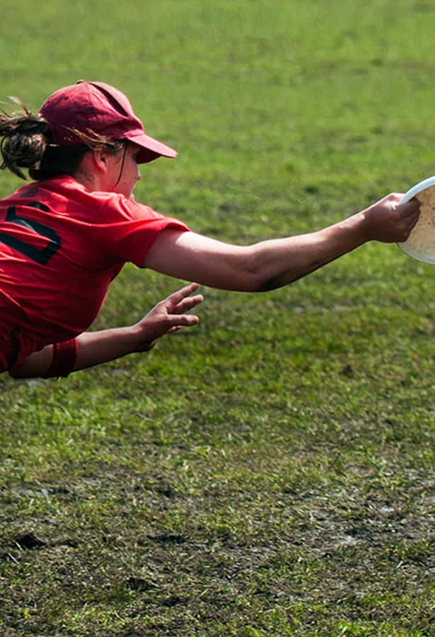 穿紅色上衣與帽子的女性運動員在Android Ultimate Frisbee背景中 壁紙 — 一位身穿紅色球衣的女運動員在草地上飛身撲接空中白色飛盤。