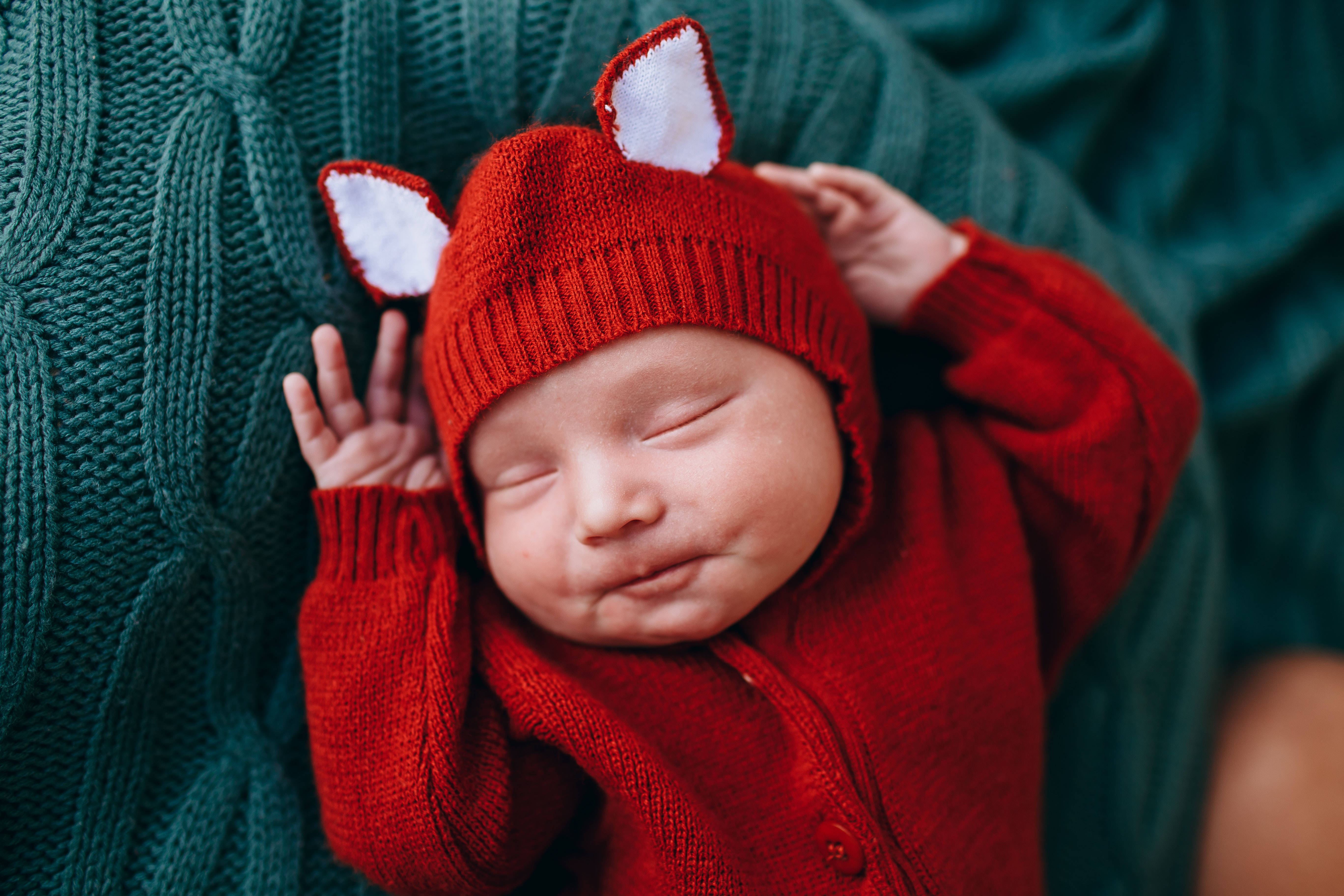 穿紅色狐狸服的超可愛嬰兒 壁紙 — 一個寧靜的新生嬰兒戴著紅色狐狸耳針織帽和配套服裝，在青綠色的電纜針織毯上安然入睡。