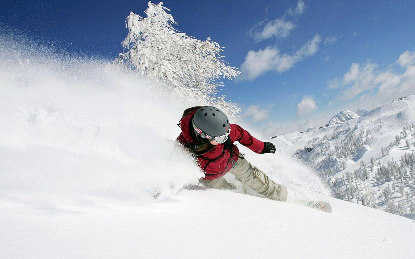 穿紅色衣服的男子雪板疾馳雪中 壁紙 — 一名身穿紅色滑雪板的滑雪者在陽光照耀的山坡上，穿過新鮮的粉雪，周圍是白雪覆蓋的樹木和藍天。