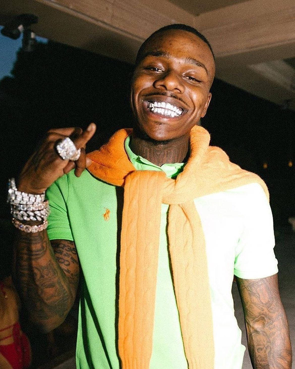 穿綠色上衣 DaBaby 手機 壁紙 — 一名微笑的男子身穿綠色襯衫和橙色圍巾，在昏暗燈光下展示鑽石戒指和鑲鑽手鐲。