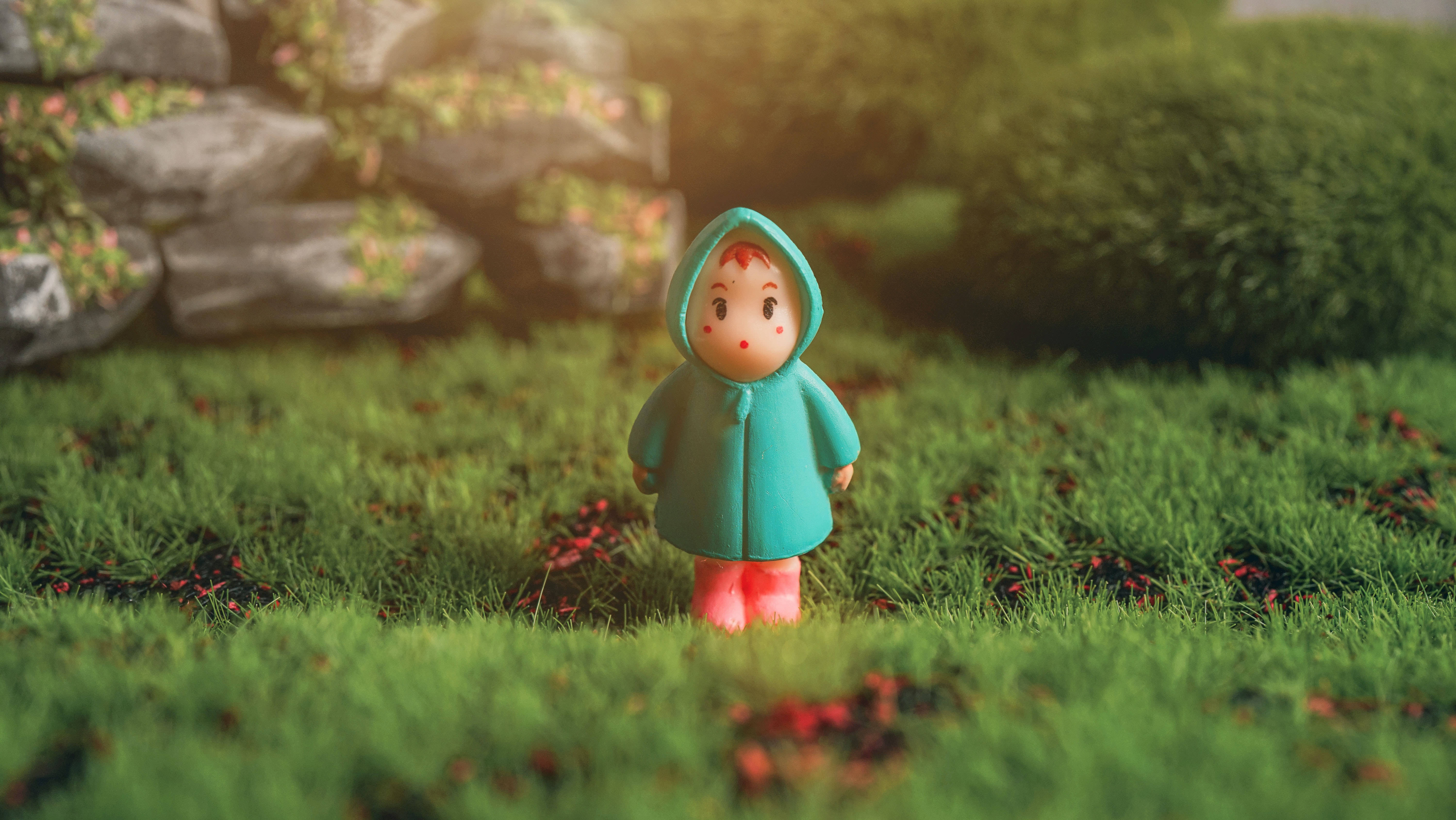 穿綠雨衣的小孩動畫桌面 壁紙 — 一個細小嘅公仔穿著青綠色連帽雨衣，獨自企喺陽光普照嘅草地上，周圍有散落嘅紅色花朵，背景係石牆。