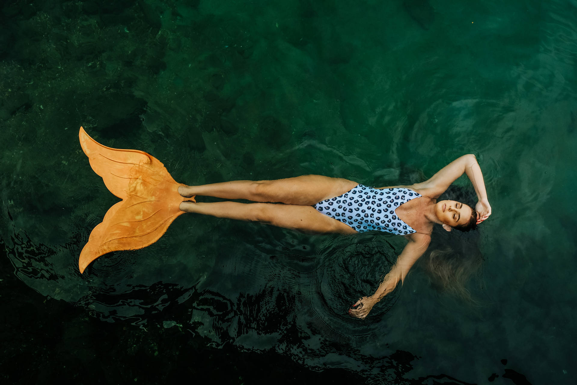 女子在泳池中仰面漂浮，穿著橙色美人魚尾巴和圖案泳衣。