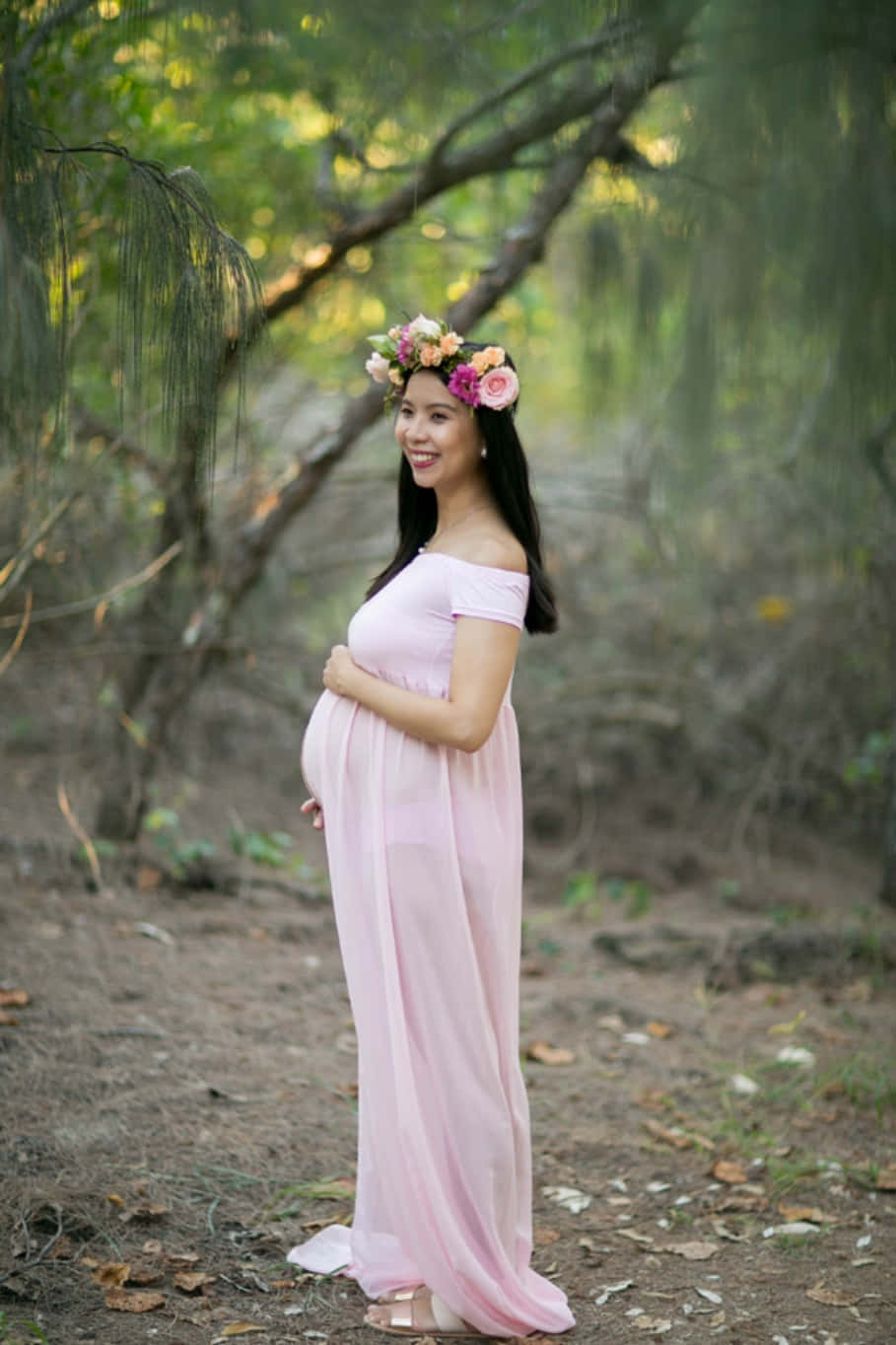 穿美麗禮服的孕婦照片 壁紙 — 一位微笑的孕婦穿著飄逸的粉紅色連衣裙，站在陽光明媚的森林中，戴著色彩繽紛的花冠。