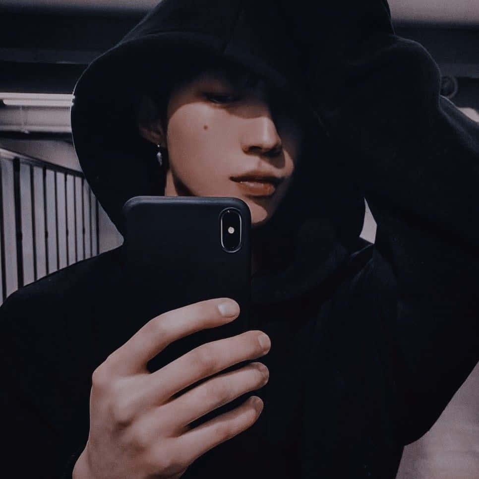 穿著 Oversized 毛衣與 Beanie 的時尚男孩 壁紙 — 一個穿著黑色連帽衫的年輕人在昏暗燈光下用智能手機對著鏡子自拍。