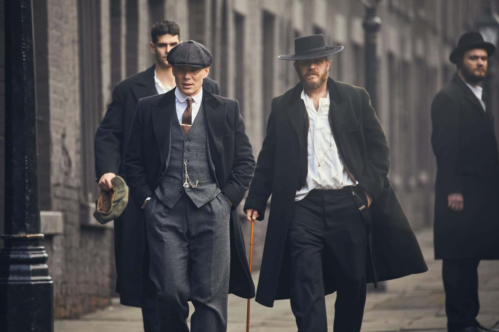 Peaky Blinders 桌布 壁紙 — 穿著復古西裝和帽子的男人自信地走在兩旁是磚砌建築的骯髒城市街道上。