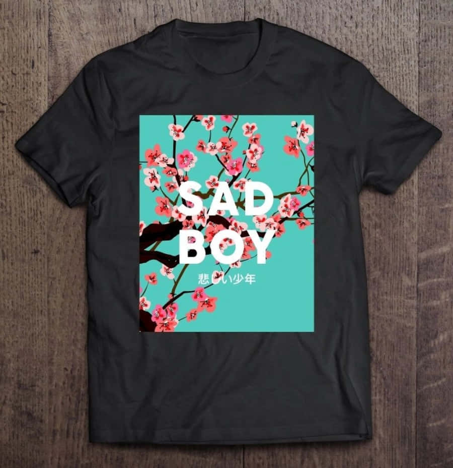 穿著時尚又舒適的T恤，展現你的風格 壁紙 — 黑色T恤，白色粗體「SAD BOY」文字和粉紅色櫻花，青色背景，放在木質表面上。