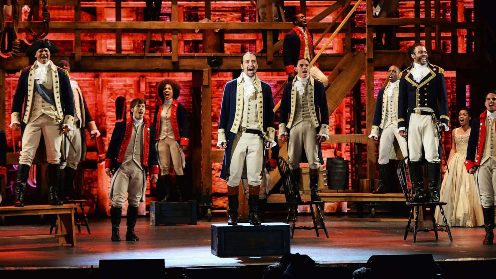Hamilton 桌布 壁紙 — 穿著殖民時期服裝的演員在紅色燈光籠罩的木製舞台上充滿活力地表演。