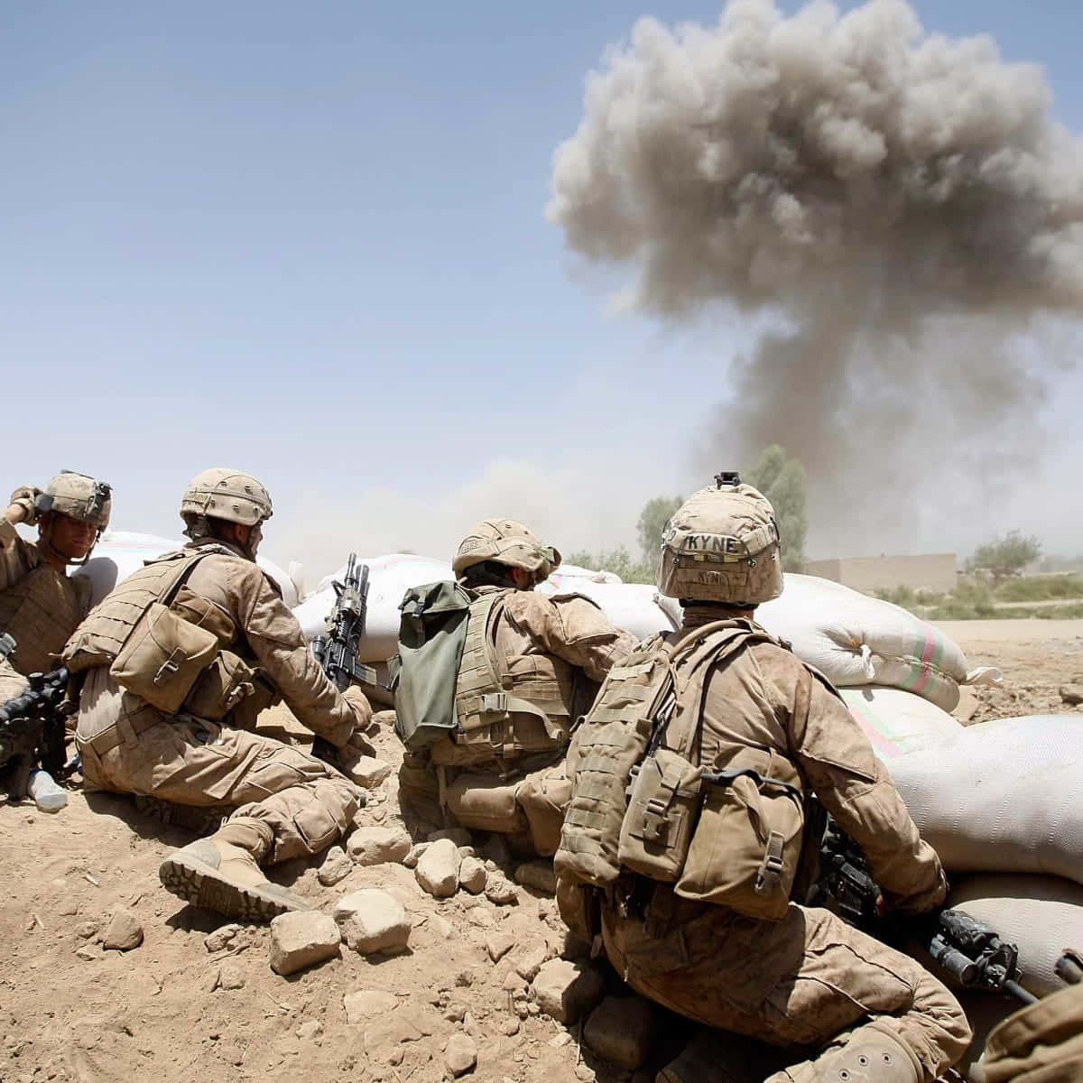 Afghanistan 桌布 壁紙 — 穿著沙漠迷彩服的士兵蹲在沙袋後面，遠處巨大的煙霧在晴朗的藍天下爆發。