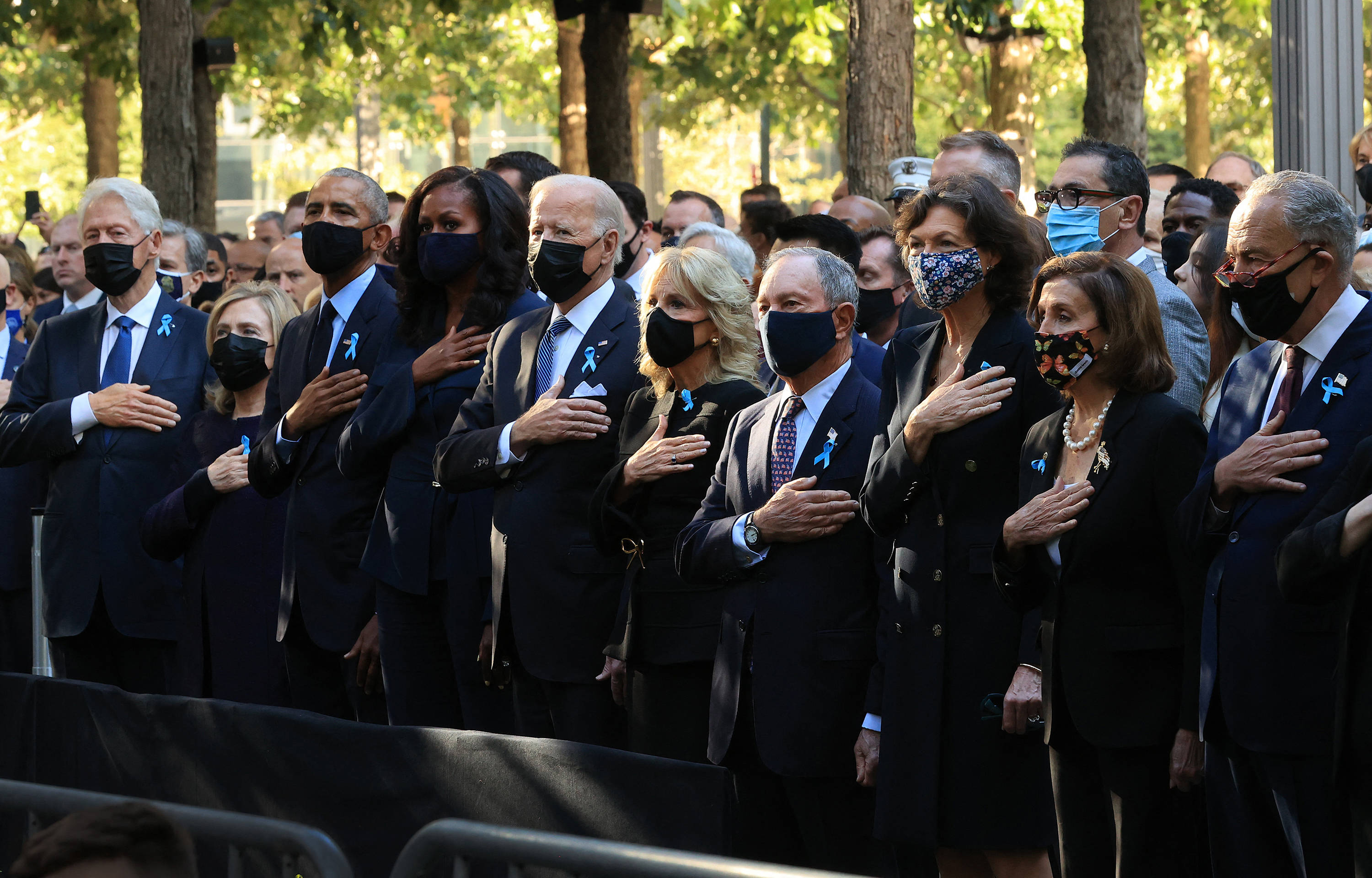 911 Memorial 桌布 壁紙 — 穿著深色西裝和口罩的政治人物，在樹木間斑駁的陽光下，莊嚴地將手放在心口上。