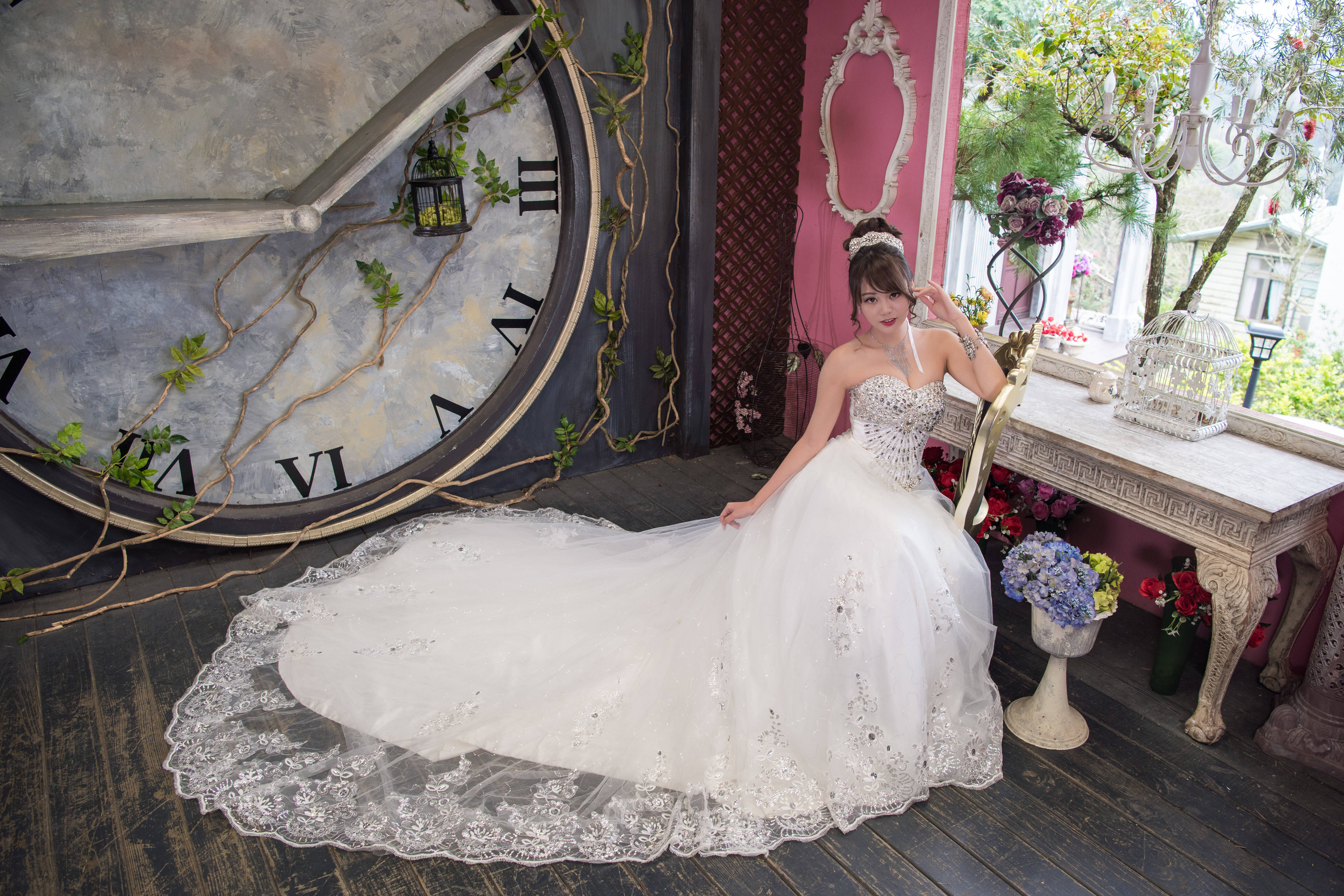 穿著白色婚紗的新娘 壁紙 — 一位身穿閃亮白色婚紗的女性優雅地坐在一個纏繞著綠色藤蔓的巨大復古時鐘旁。