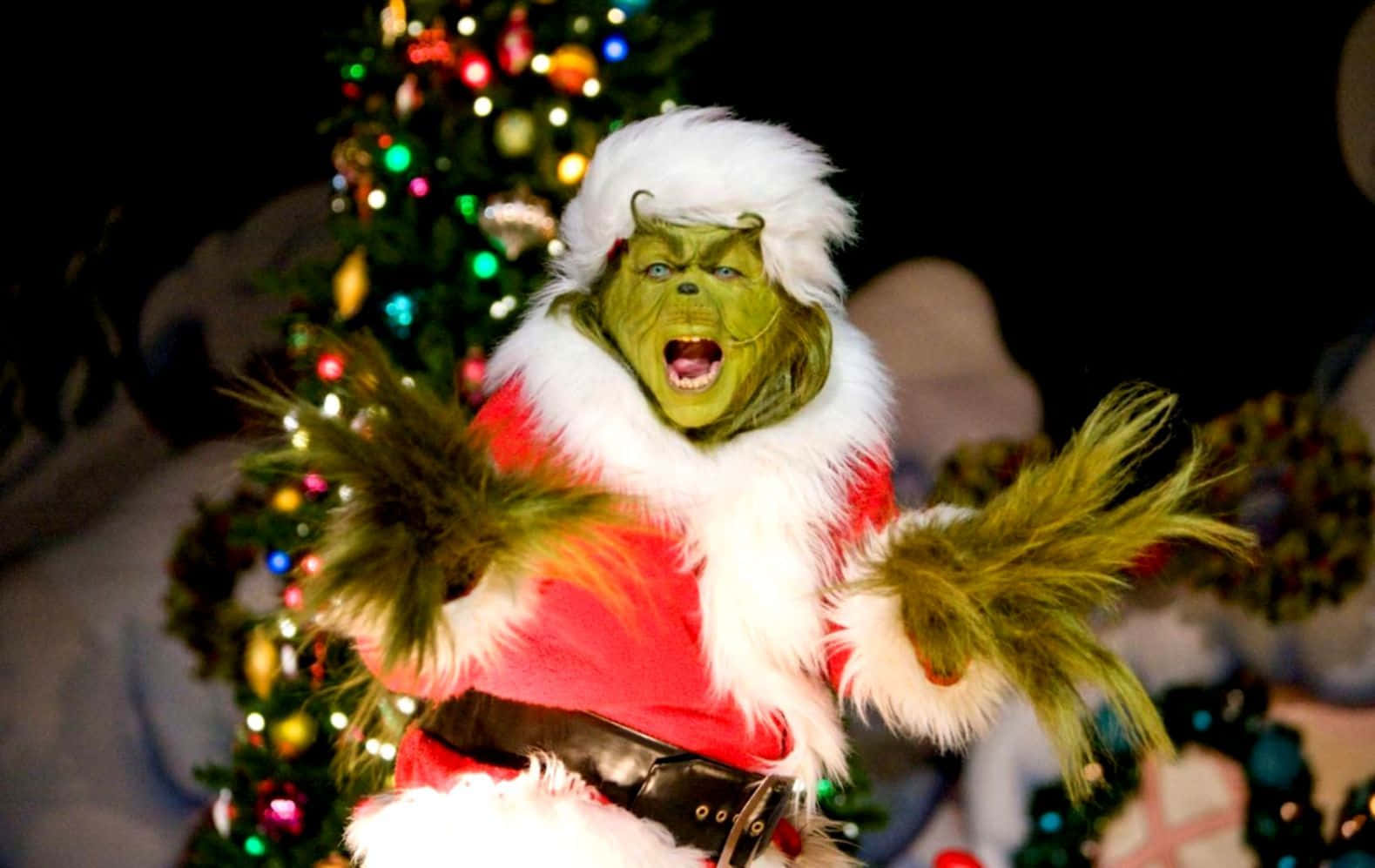 Christmas Movie 桌布 壁紙 — 穿著節日紅色套裝、白色毛邊和綠色毛手套的鬼靈精站在明亮燈飾的聖誕樹前。