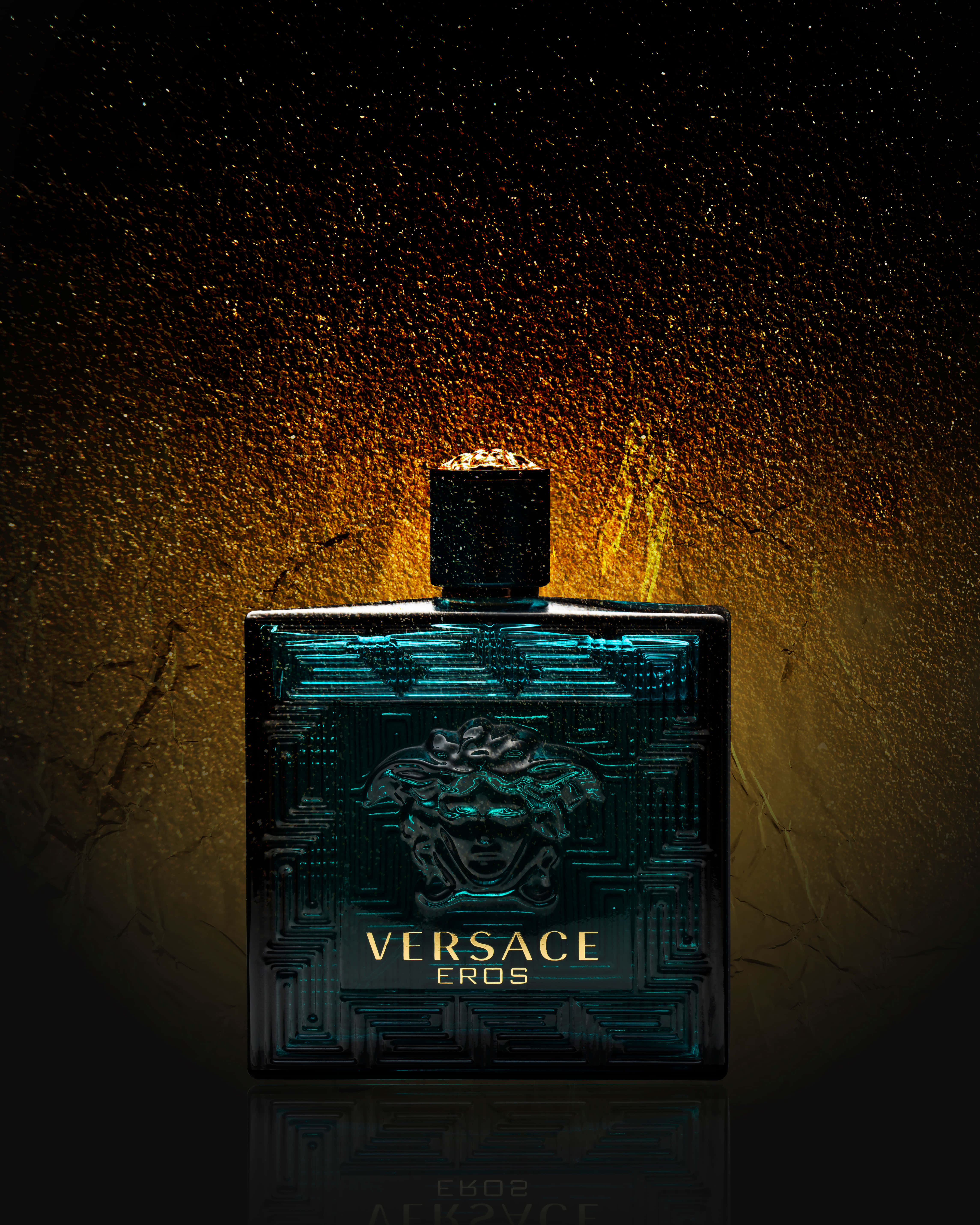 穿著范思哲，展現優雅氣質 壁紙 — 時尚的 Versace Eros 香水瓶在質感金色背景上閃爍著青綠色的光芒，背景上點綴著閃閃發光的顆粒。