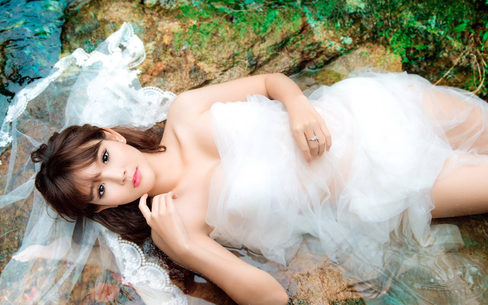 穿著華麗婚紗的新娘 壁紙 — 一位身穿飄逸白色婚紗和頭紗的女性，在清澈的水中靠著長滿苔蘚的岩石，以柔和的表情向上凝視。