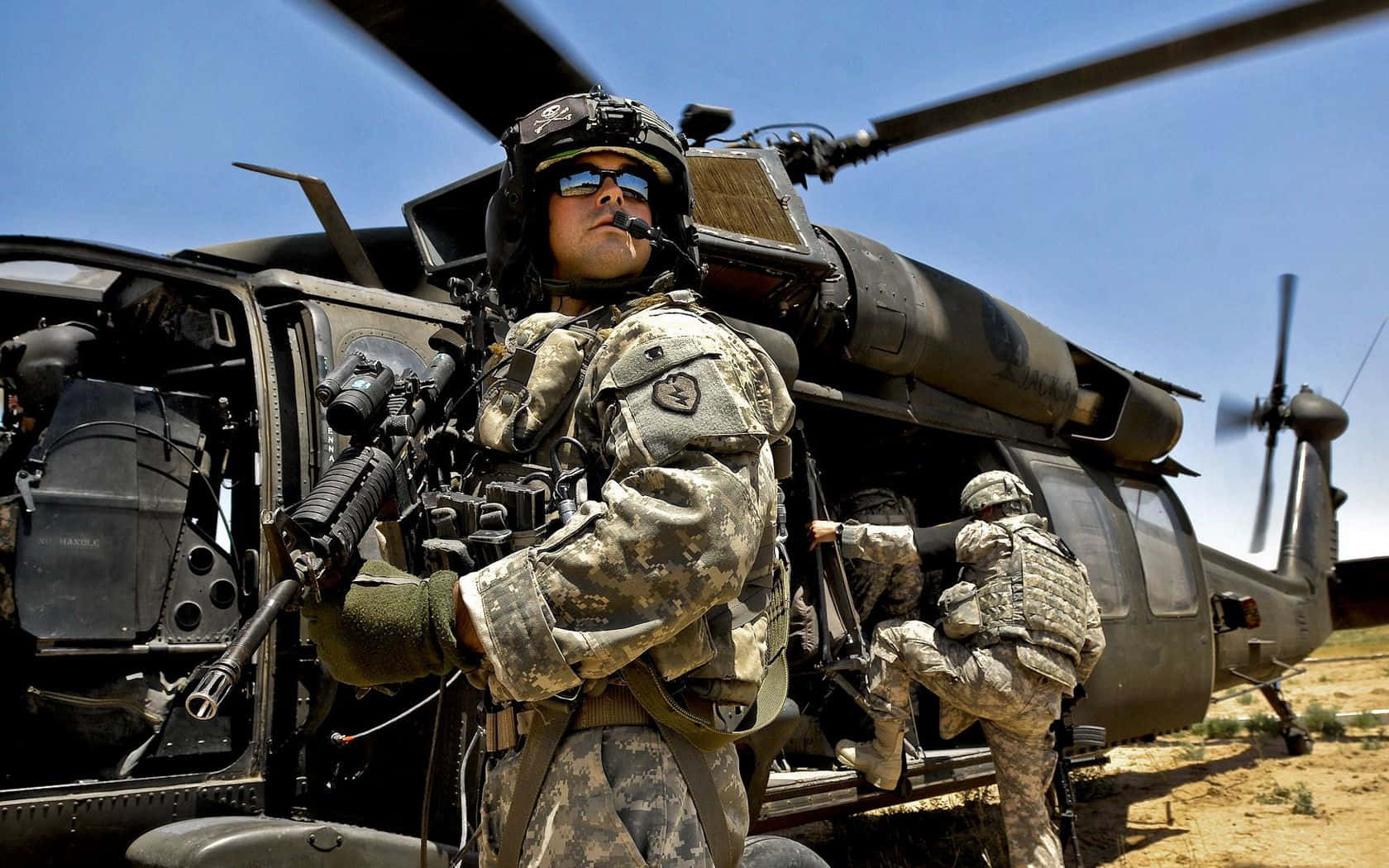 Army 桌布 壁紙 — 穿著迷彩裝備的士兵在沙漠環境中，在晴朗的藍天下，警覺地站在一架黑色軍用直升機旁邊。