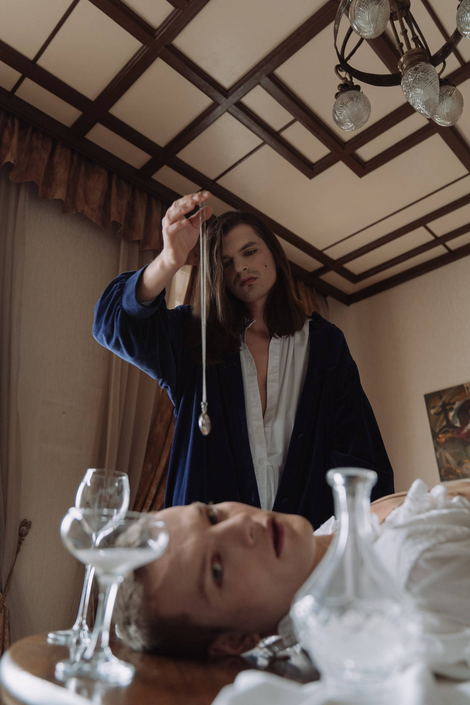 穿藍夾克女子進行催眠 壁紙 — 一個穿著藍色長袍的人，在一個光線昏暗、有吊燈的房間裡，將一個鐘擺懸在躺在桌子上方的另一個人上方。