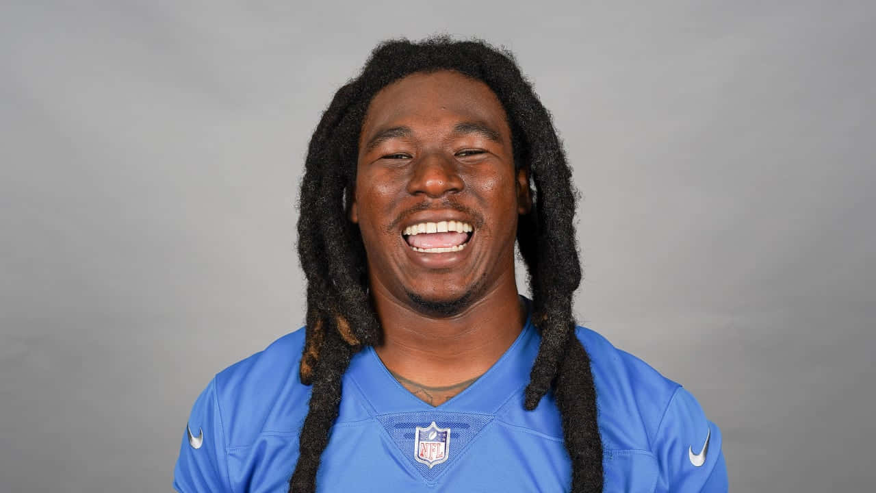 穿藍色球衣微笑的足球運動員 壁紙 — 一位快樂的NFL球員留著長辮子，穿著藍色獅子隊球衣，在純灰色工作室背景前開懷大笑。