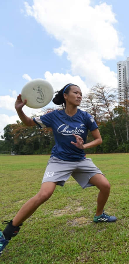 穿藍色運動服亞洲女運動員 Android Ultimate Frisbee 背景 壁紙 — 一位專注的女運動員穿著藍色球衣，在陽光明媚的草地上擲出白色飛盤，背景有樹木和高樓大廈。