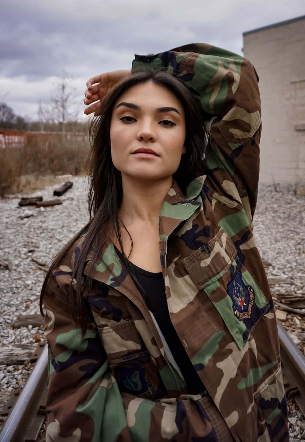 穿軍裝的女性擺姿勢 壁紙 — 一名身穿迷彩外套的女子自信地站在鐵路軌道上，背景是陰天和光禿禿的樹木。