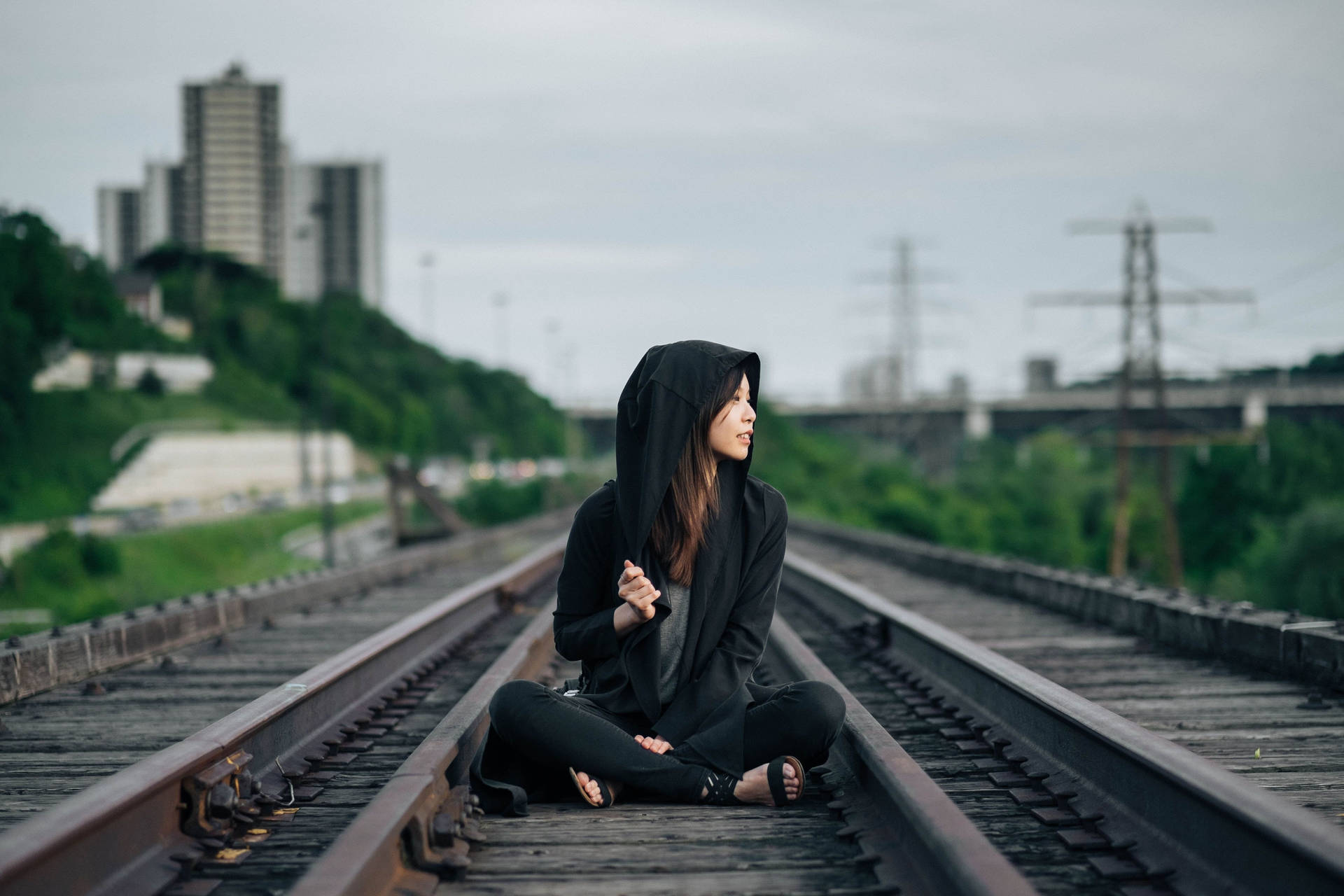 穿著黑色連帽衫的女性坐在火車軌道上，在城市環境中望向遠方