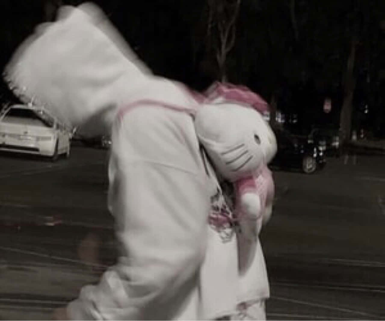 穿著白色連帽衫的人夜晚在停車場中拿著粉紅色Hello Kitty毛絨玩具。