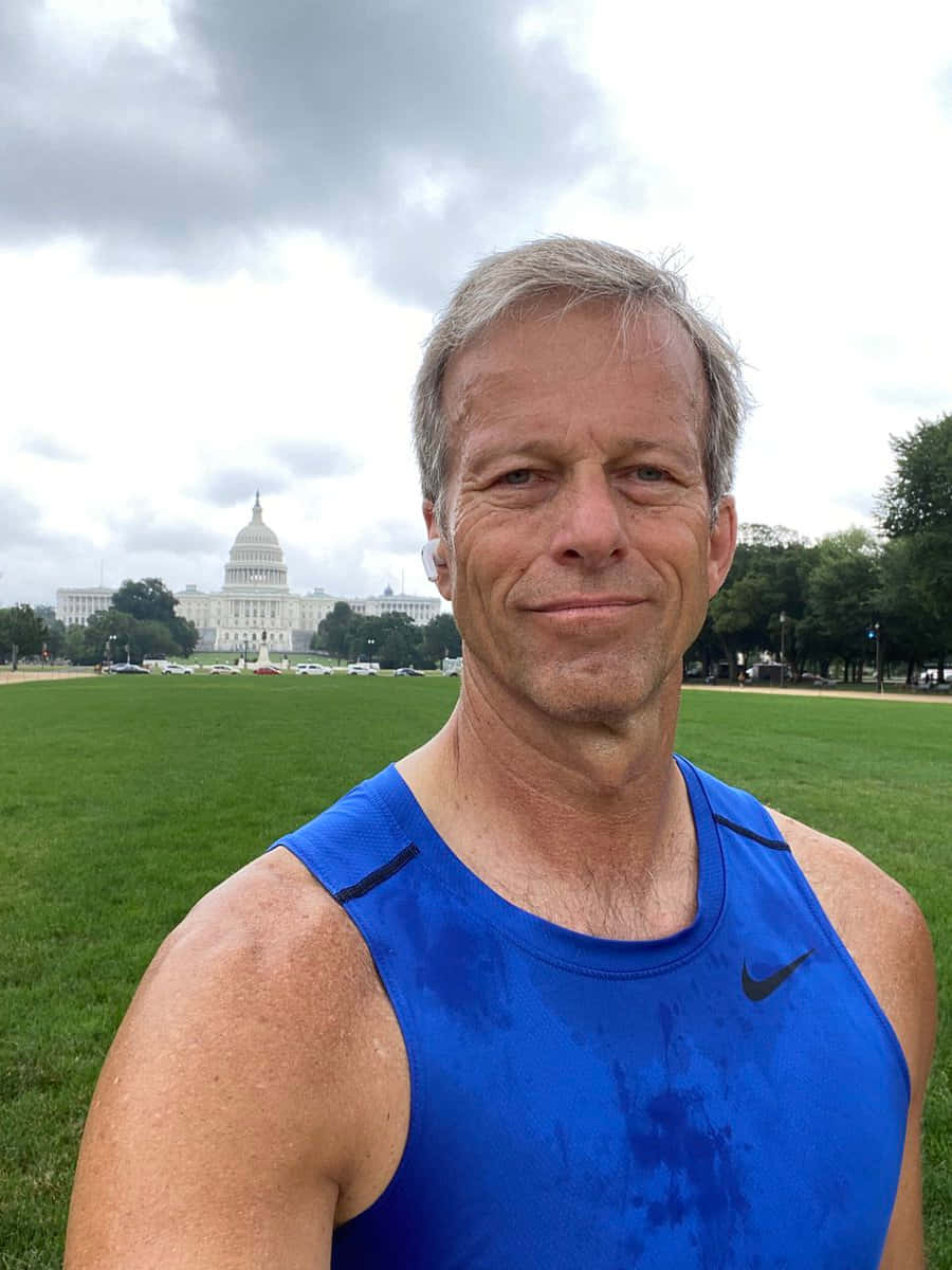 穿運動服的參議員約翰·桑恩 壁紙 — 一名身穿藍色運動背心的男士在陰天微笑著望向鏡頭，身後可見美國國會大廈。
