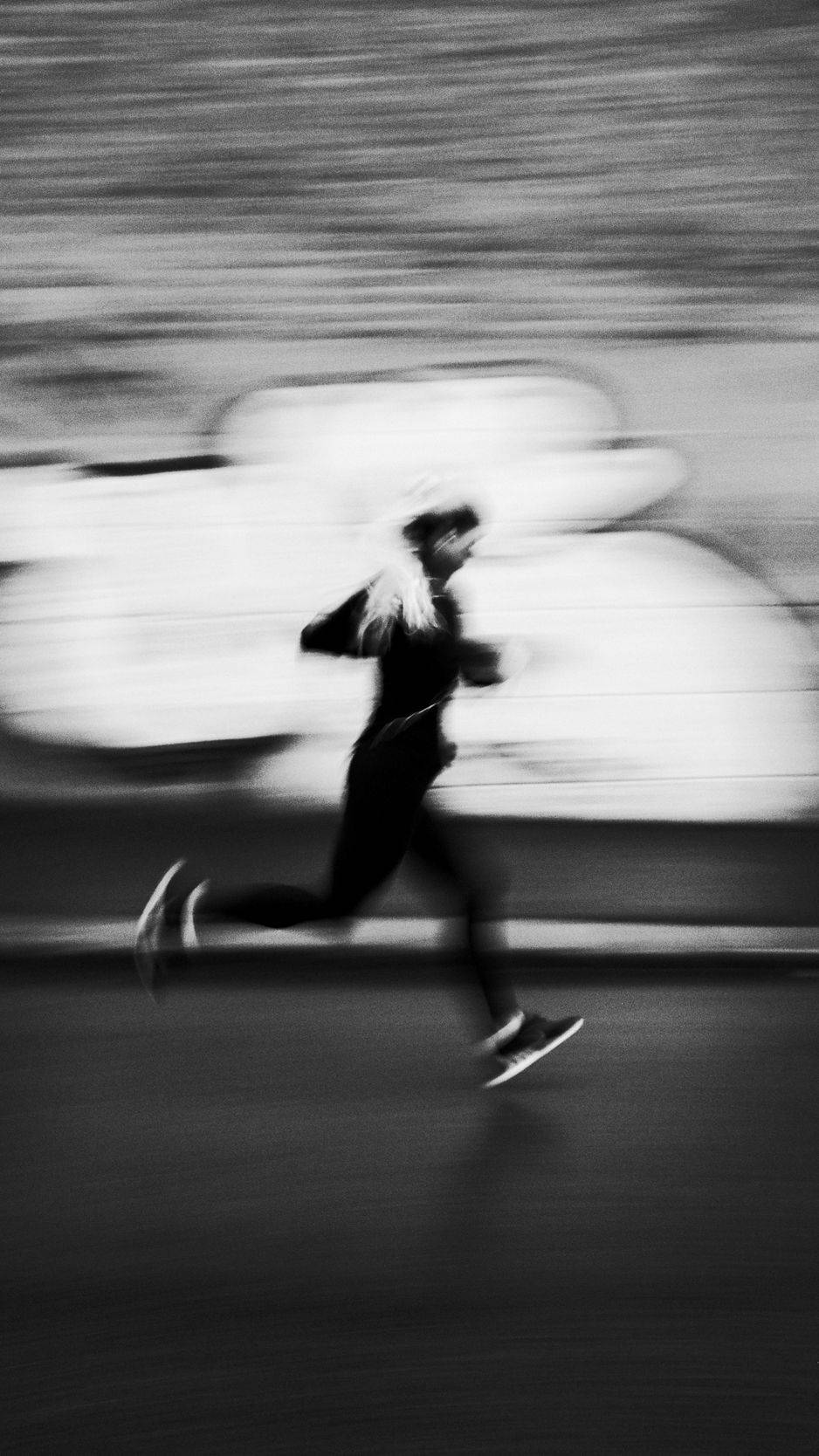 穿運動服跑步的男子 iPhone 體育壁紙 壁紙 — 以單色調捕捉嘅跑步者，喺有條紋嘅城市背景下呈現戲劇性嘅動態模糊效果。