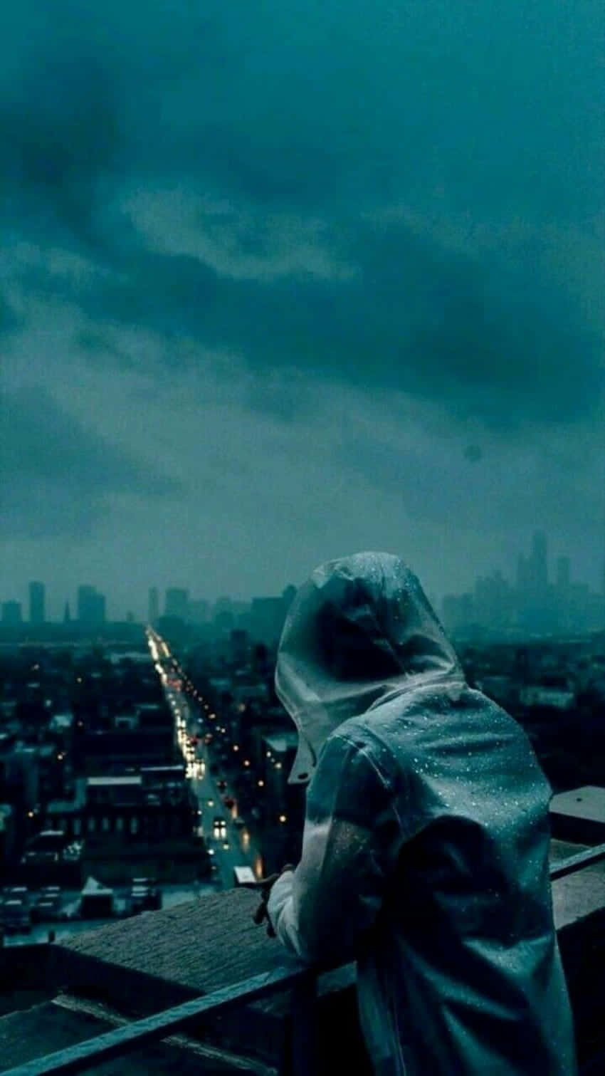 穿雨衣的冷靜憂傷男孩 壁紙 — 一個孤獨的戴帽人影站在屋頂上，俯瞰著在憂鬱的青色天空下被雨水浸濕的城市景觀。