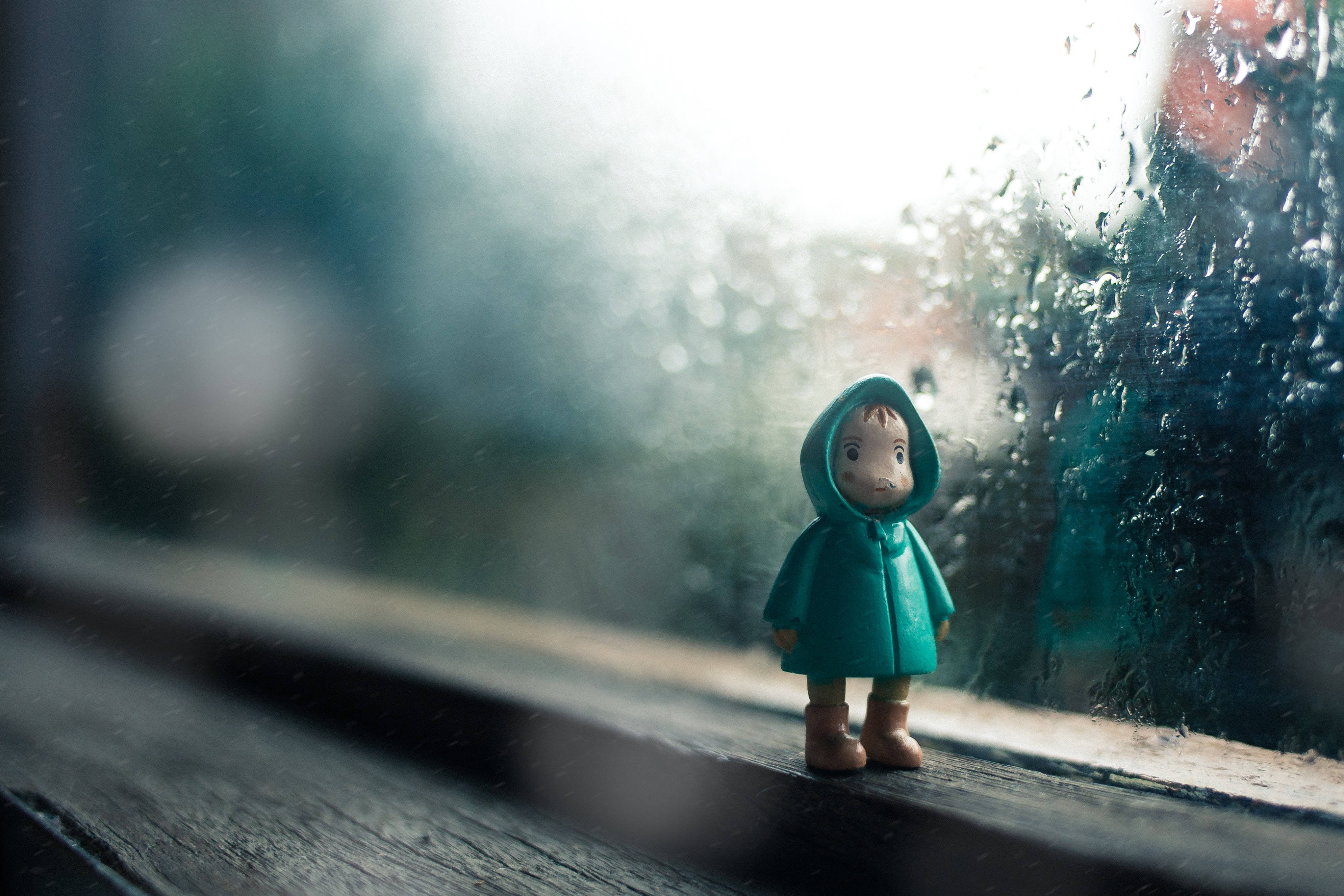 穿雨衣的小孩動畫桌面壁紙 壁紙 — 一個穿著青綠色雨衣的小人偶獨自站在窗台上，凝視著窗外雨濛濛、模糊的世界。
