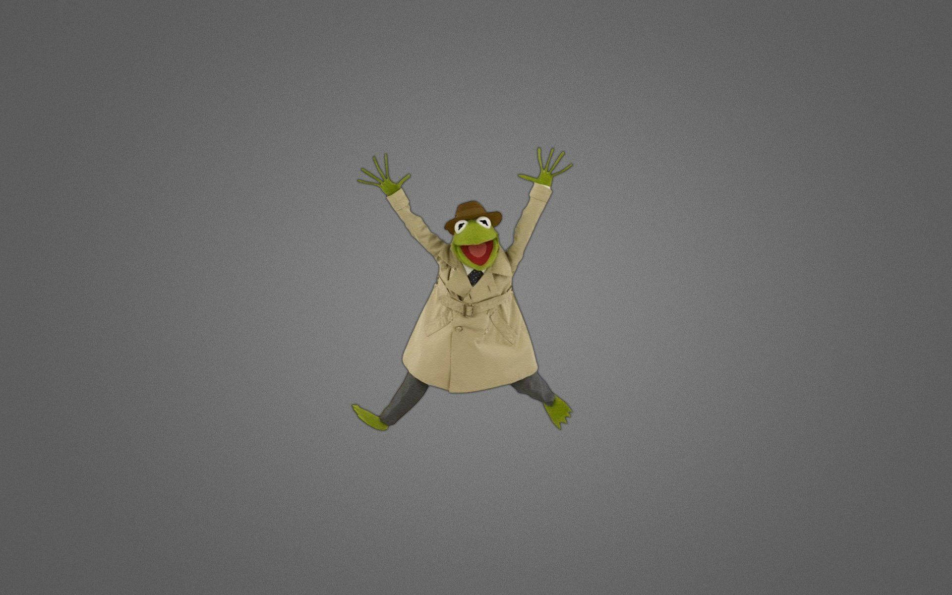 穿風衣的Kermit青蛙 壁紙 — Kermit the Frog 穿著米色乾濕褸和軟呢帽，在純灰色背景上舉起雙臂歡快地跳躍。