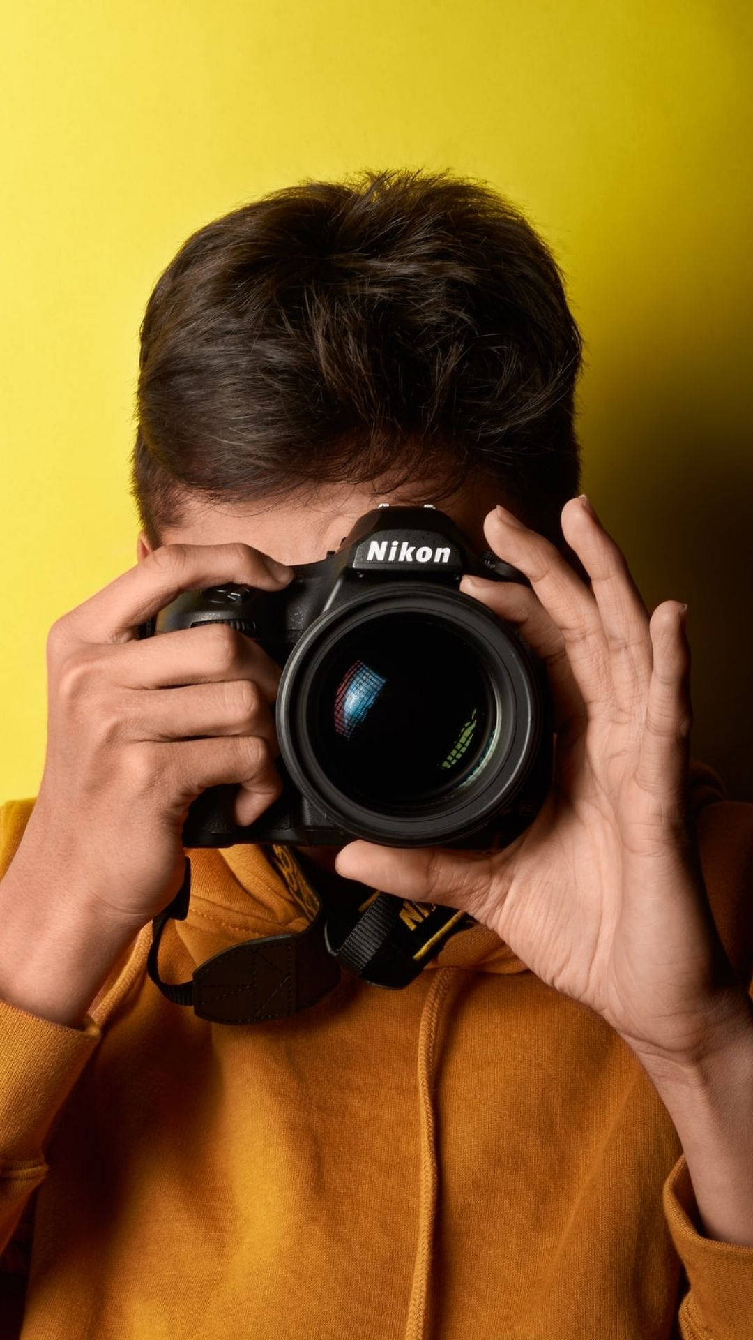 穿黃色外套的攝影師 壁紙 — 一名身穿橙色連帽衫的人將黑色Nikon DSLR相機舉到臉前，背景為鮮豔的黃色。