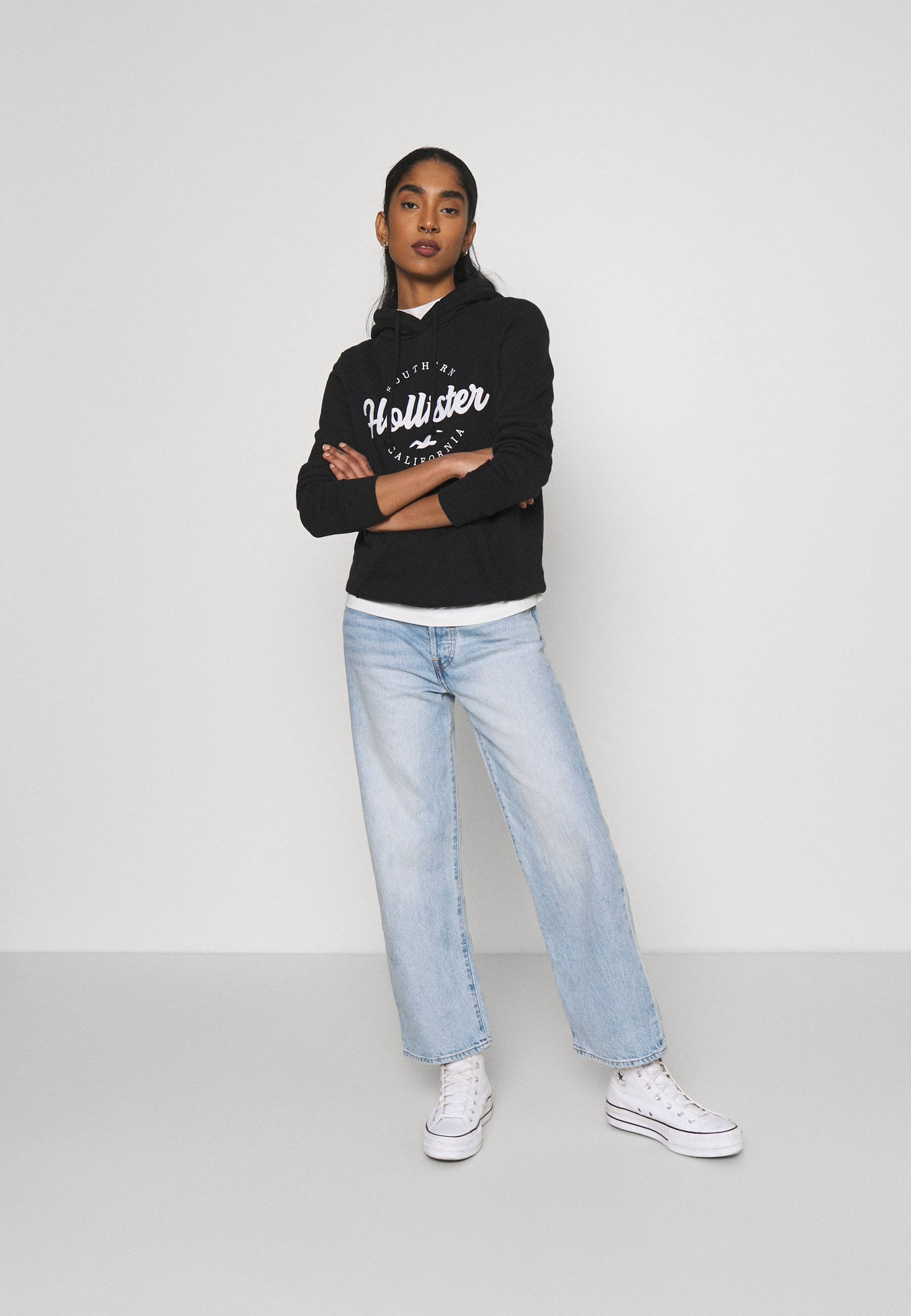 穿黑色Hollister連帽外套的模特 壁紙 — 一位年輕女性自信地交叉雙臂站立，穿著黑色Hollister連帽衫、淺藍色牛仔褲和白色運動鞋，背景是純白色攝影棚。