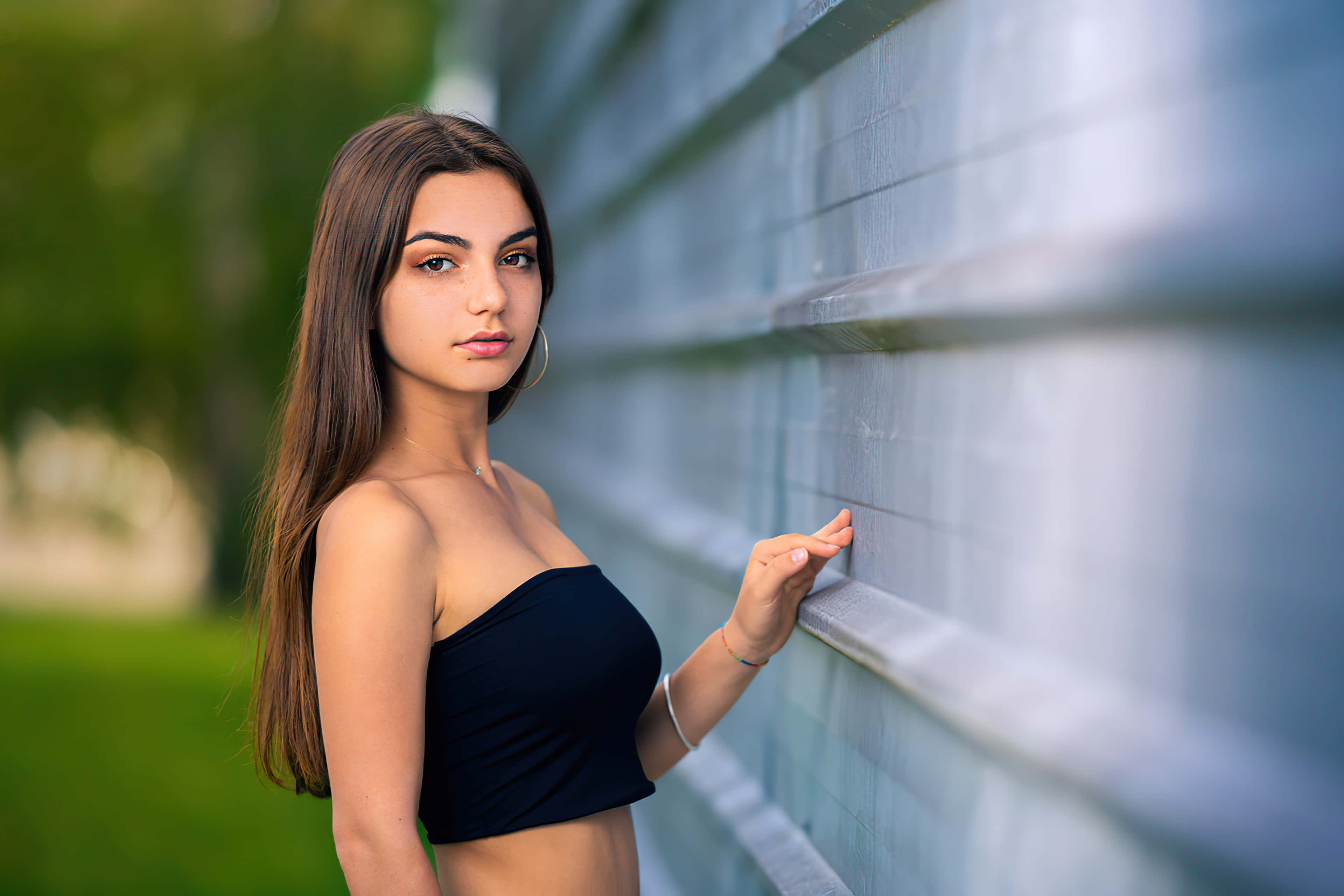 穿黑色上衣的少女 壁紙 — 一位身穿黑色上衣的年輕女性靠著紋理灰色牆壁，背景有綠色樹葉柔和模糊。