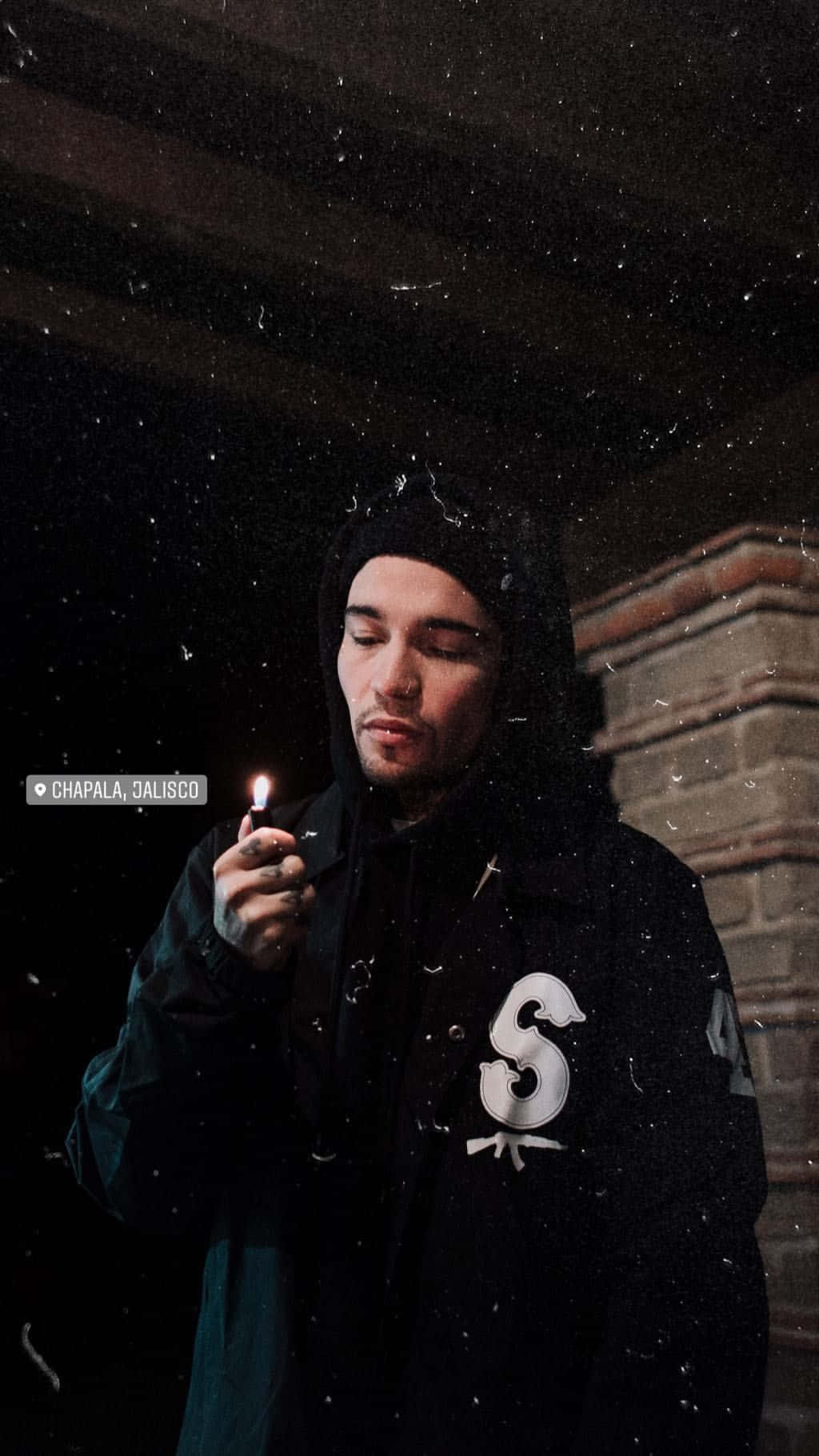 穿黑色連帽衫的帥哥 壁紙 — 一個穿著黑色連帽衫的男人在雪夜背景下，手持點燃的打火機，背景是磚砌建築。