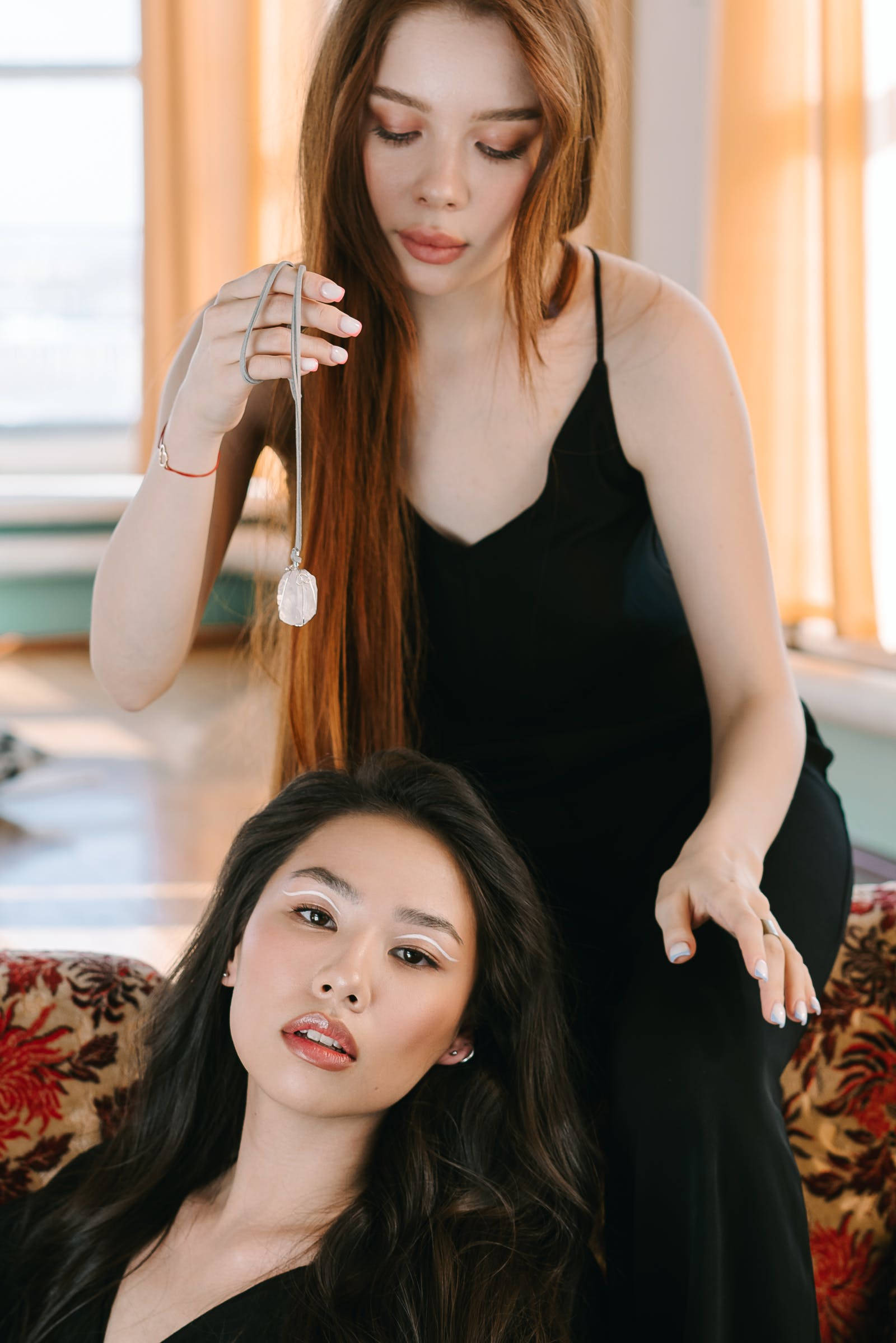 穿黑裙女子進行催眠 壁紙 — 兩位身穿黑色連衣裙的女性，一位坐在花卉沙發上，另一位站著手持水晶吊墜，室內場景。