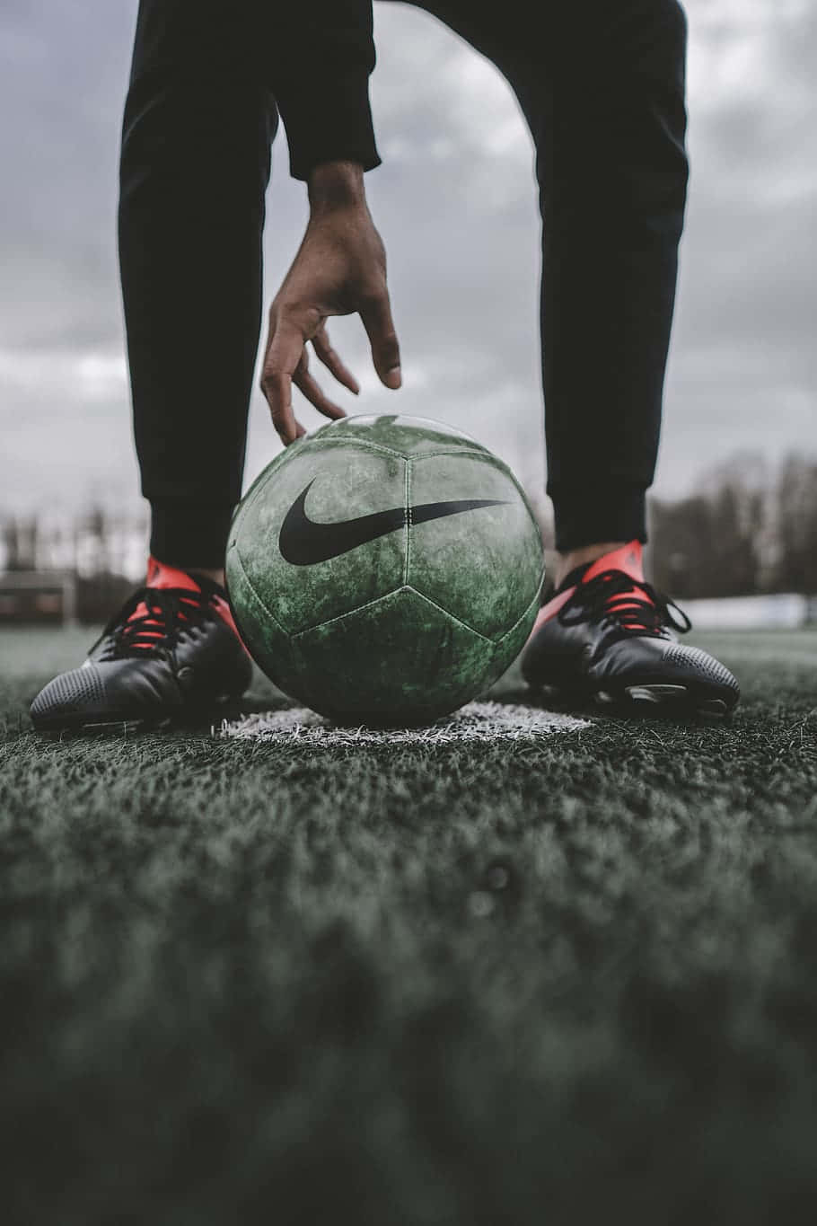 穿黑褲的人在場地上撿起帶Nike標誌的足球 壁紙 — 球員雙腳穿著黑色釘鞋配紅色鞋帶的特寫，準備在陰天的人造草皮上踢一個風化的綠色Nike足球。