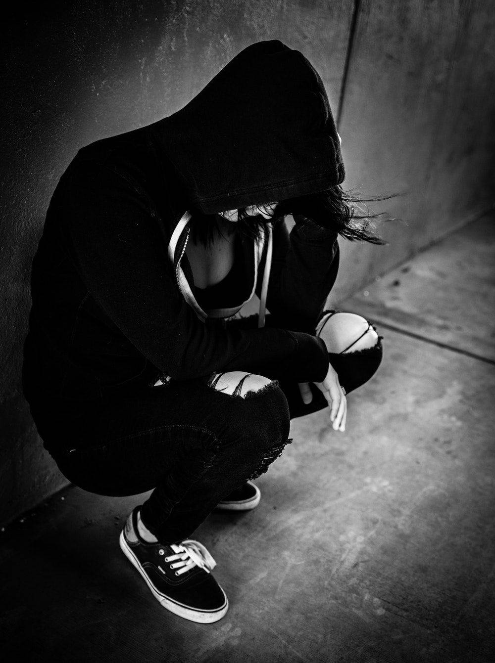 穿黑連帽衫的女孩 壁紙 — 一個穿著黑色連帽衫的人蹲在混凝土牆邊，背景是單色調的簡樸場景。