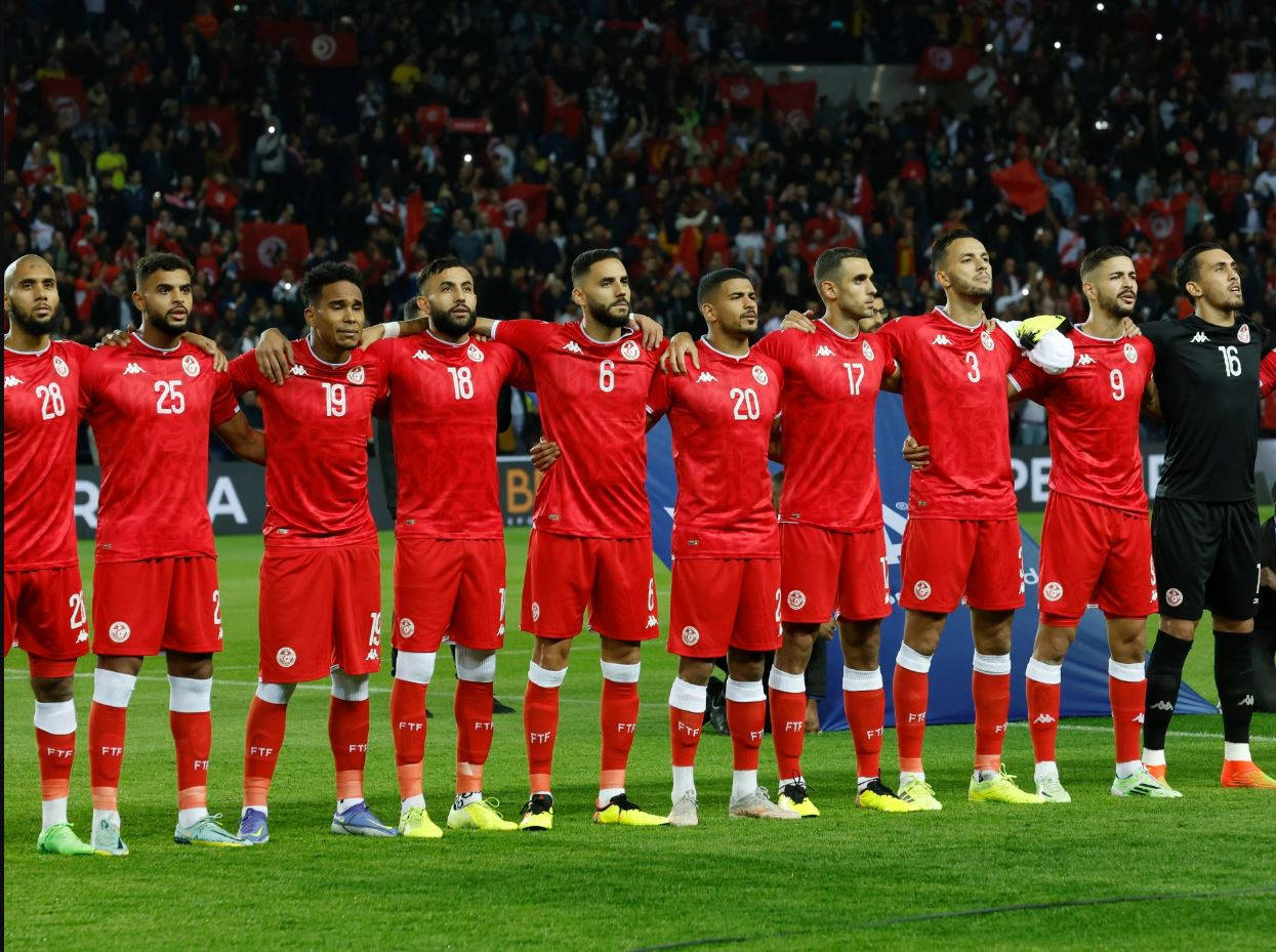 Tunisia 桌布 壁紙 — 突尼斯足球隊身穿紅色球衣，在體育場燈光下肩並肩站在充滿活力的綠色球場上，背景有球迷揮舞旗幟。