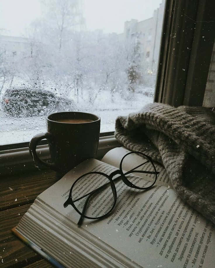 Winter Photography 桌布 壁紙 — 窗台上放著一杯冒熱氣的馬克杯和一本打開的書，窗外雪花飄落，營造出舒適的冬日場景。