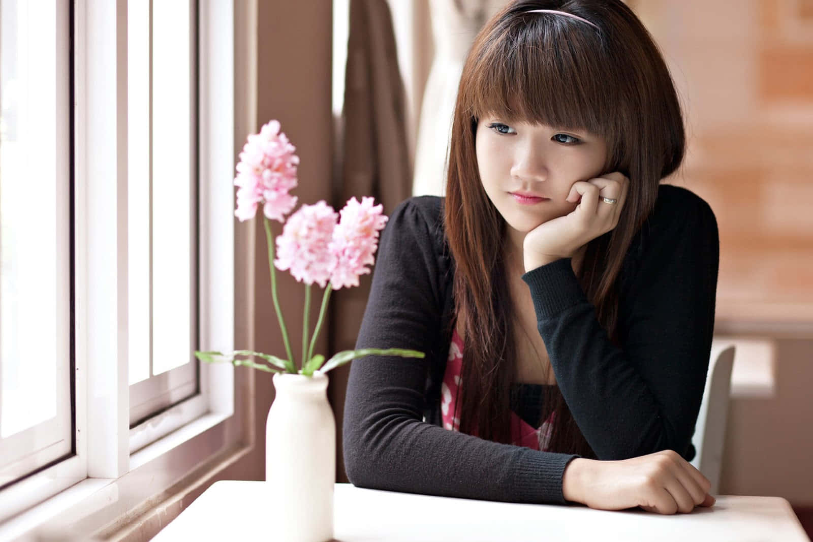 窗邊的可愛韓國女孩 壁紙 — 一位有瀏海的年輕女子在陽光照射的窗邊，將下巴擱在手上，旁邊是白色花瓶中的粉紅色花朵。