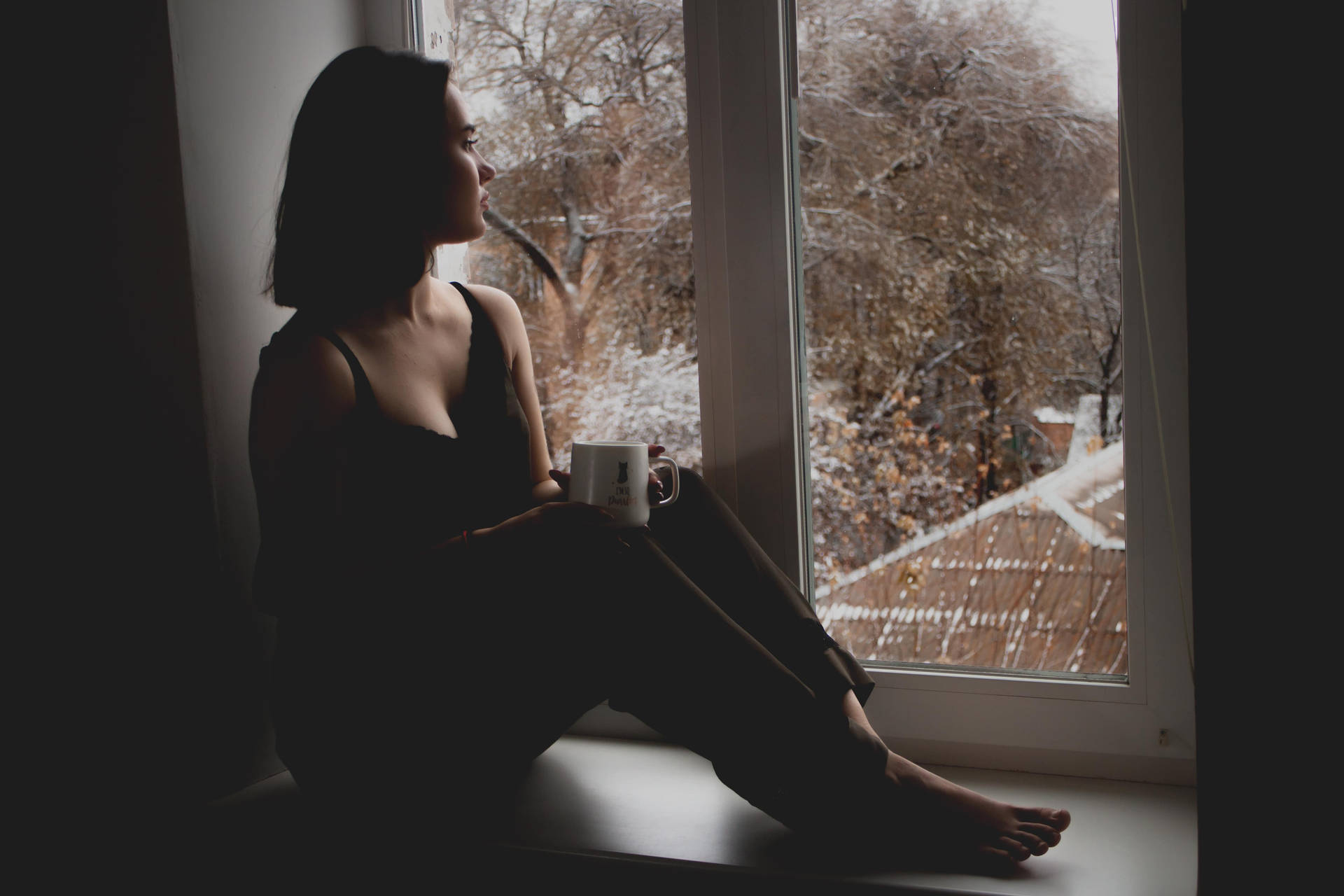 剪影中的女人在窗邊拿著杯子，看著被雪覆蓋的樹木