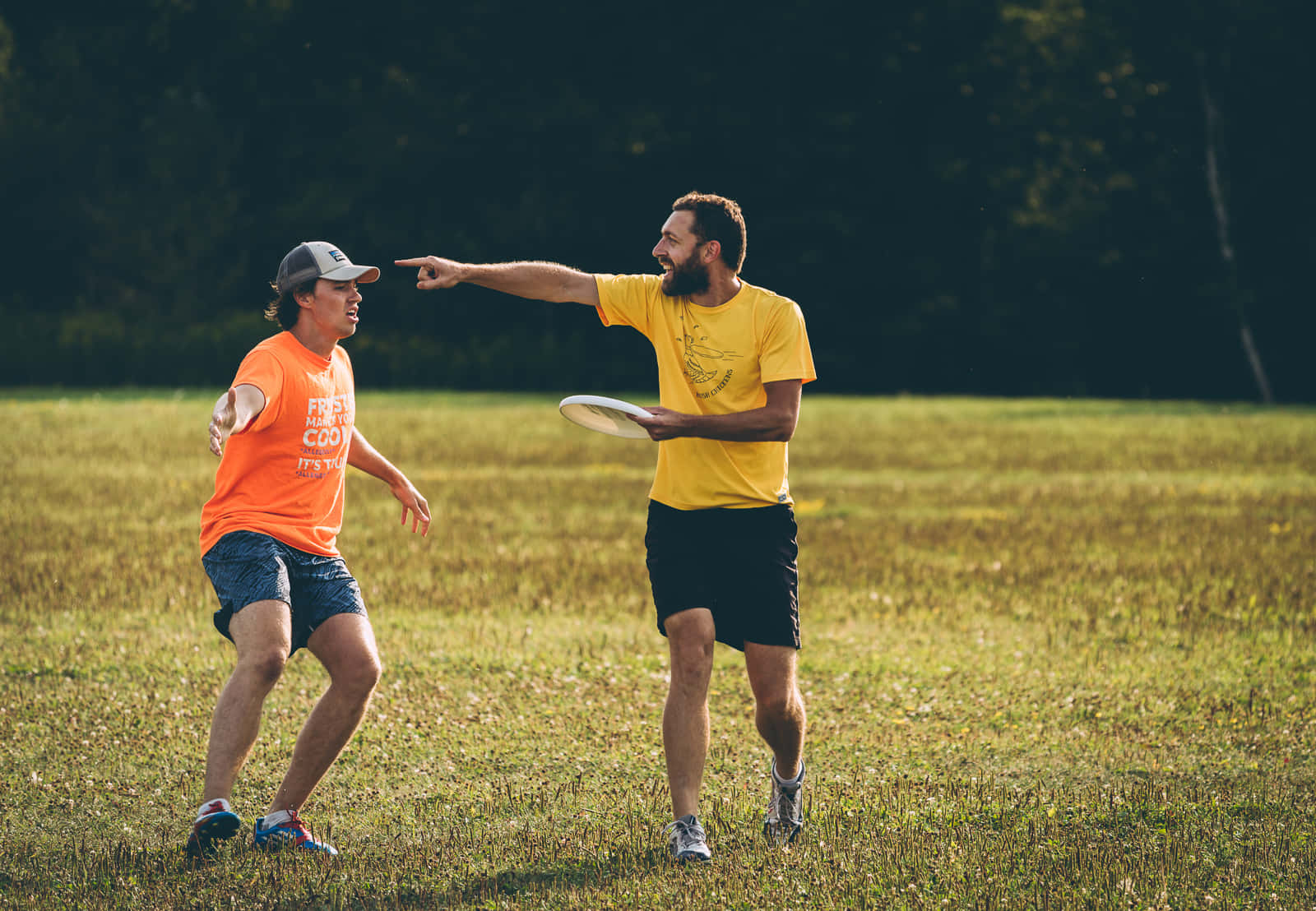 競技 Ultimate Frisbee 空中動作 壁紙 — 兩名身穿亮色襯衫的男子在陽光明媚的草地上玩飛盤，背景有樹木。