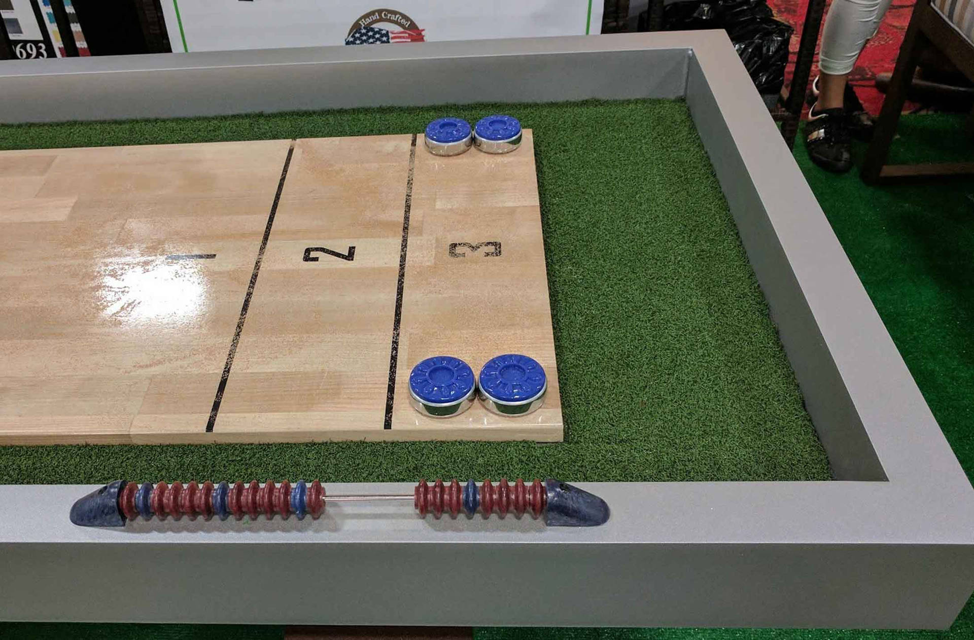 競技樂趣與堅固戶外 Shuffleboard 桌 壁紙 — 一張沙狐球桌的特寫，上面有藍色冰球和一支紅藍條紋的球桿，放在綠色人造草皮上。