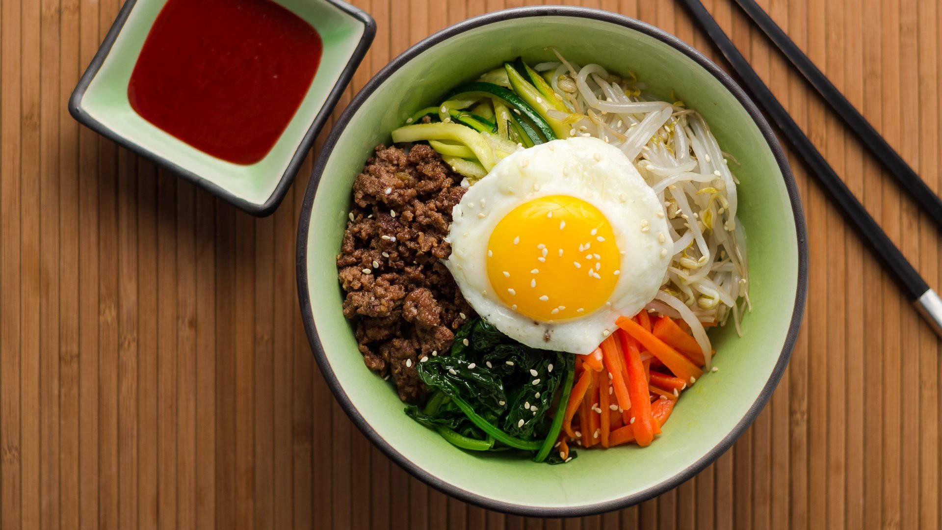 竹席上的韓式拌飯 壁紙 — 一碗色彩繽紛的韓國石鍋拌飯，上面放著太陽蛋和五顏六色的蔬菜，放在木桌上。