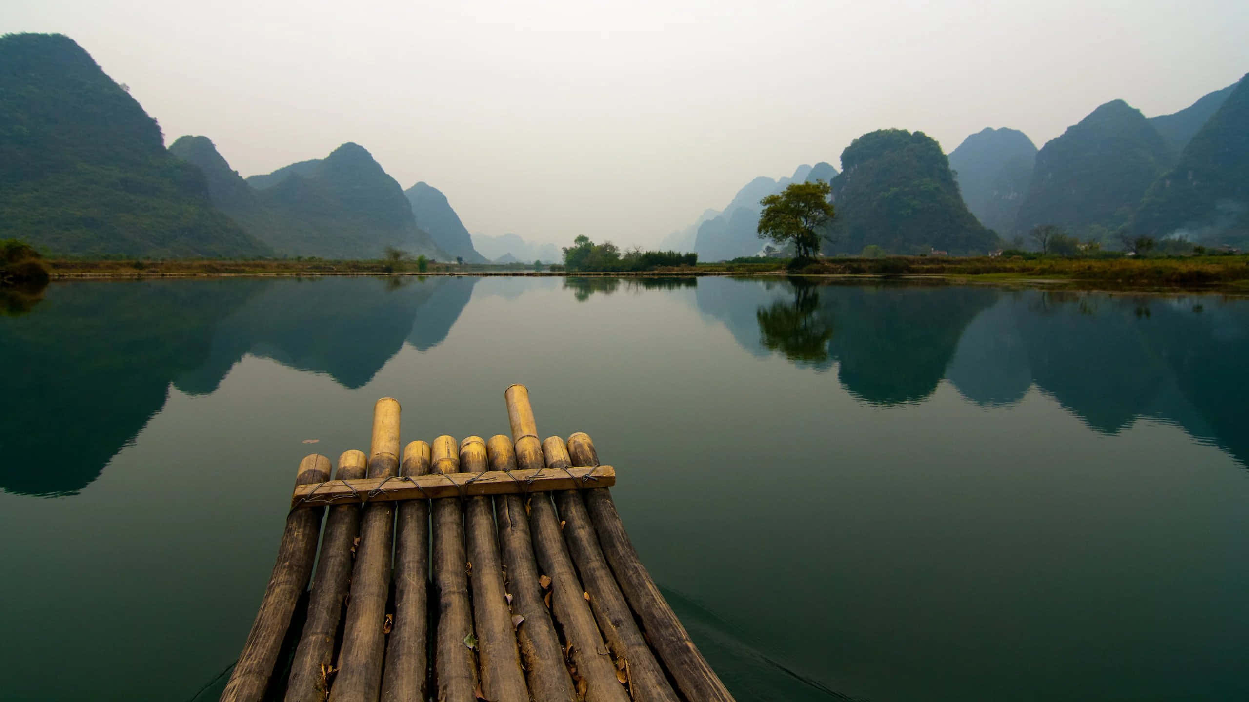 Vietnam 桌布 壁紙 — 竹筏漂浮在平靜的河面上，映照著多雲天空下霧氣繚繞的喀斯特山脈。